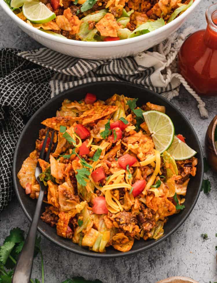 A plated serving of Dorito taco salad in a black bowl, garnished with fresh cilantro, diced tomatoes, shredded cheddar cheese, and lime wedges. The bowl sits on a textured gray surface, with a larger serving bowl in the background, a bottle of taco sauce, and fresh ingredients scattered around, creating a vibrant and appetizing presentation.