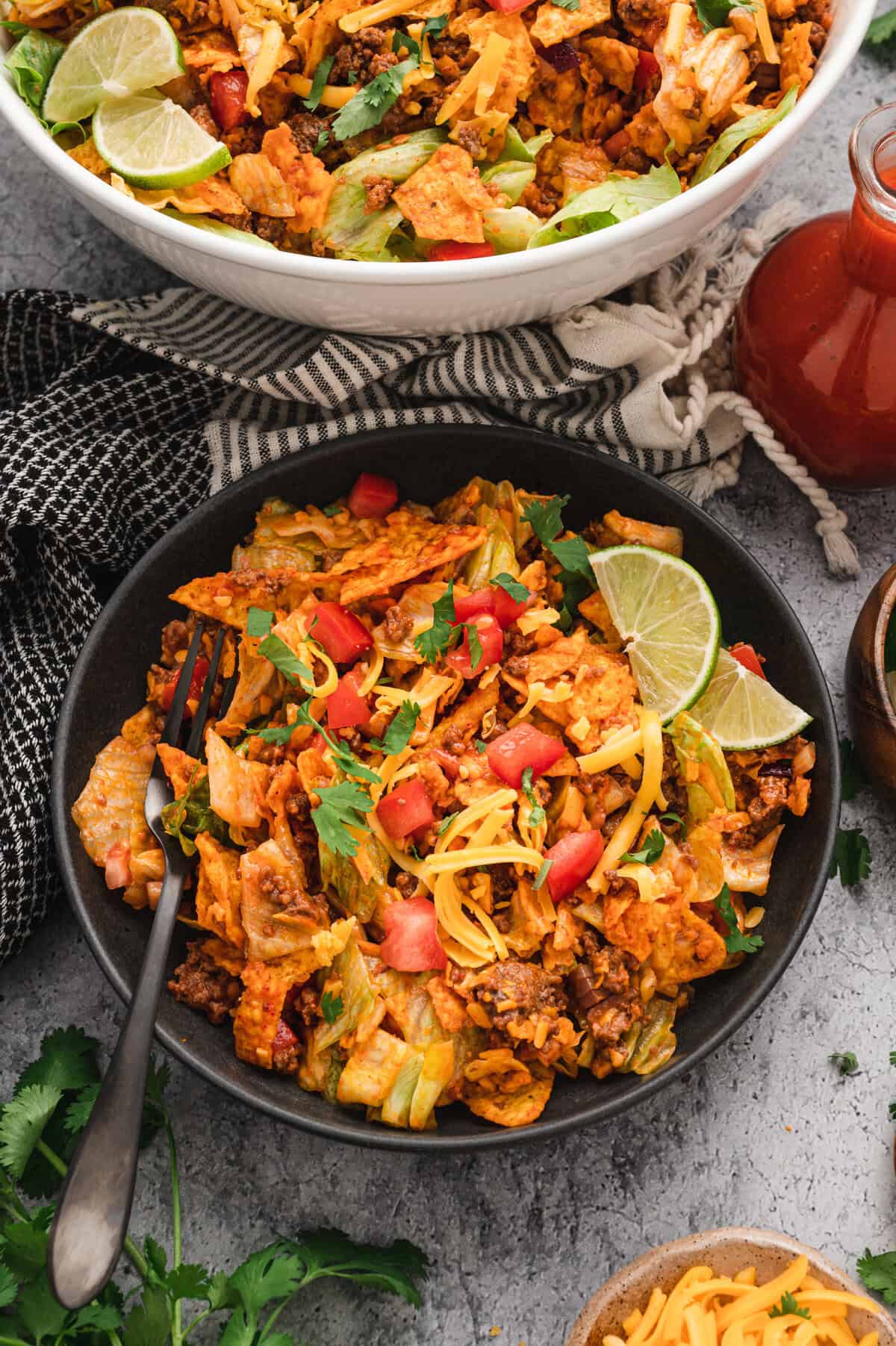 A plated serving of Dorito taco salad in a black bowl, garnished with fresh cilantro, diced tomatoes, shredded cheddar cheese, and lime wedges. The bowl sits on a textured gray surface, with a larger serving bowl in the background, a bottle of taco sauce, and fresh ingredients scattered around, creating a vibrant and appetizing presentation.