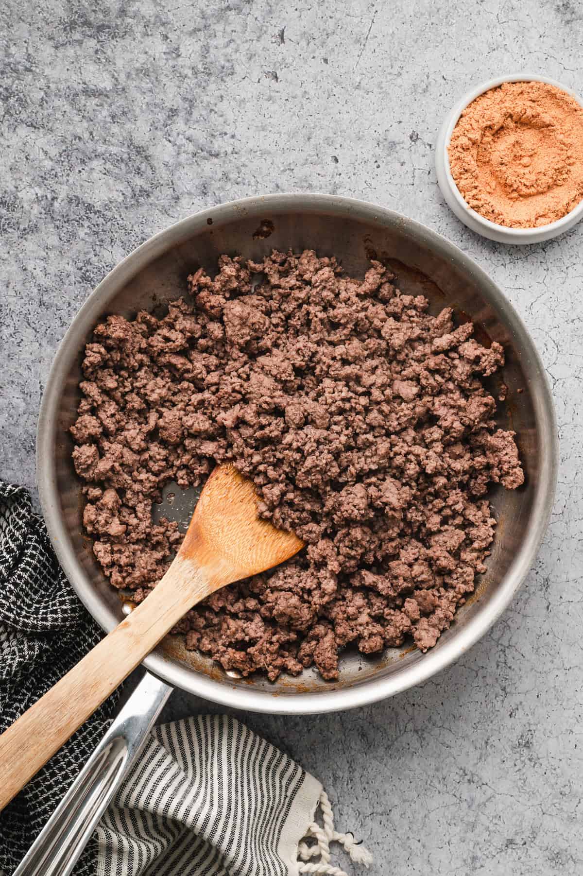 A stainless steel pan filled with cooked ground beef sits on a textured grey surface. A wooden spoon rests in the pan, partially submerged in the beef. A small white bowl containing an orange-colored seasoning is placed nearby. Black and white striped kitchen towels are draped around the scene.