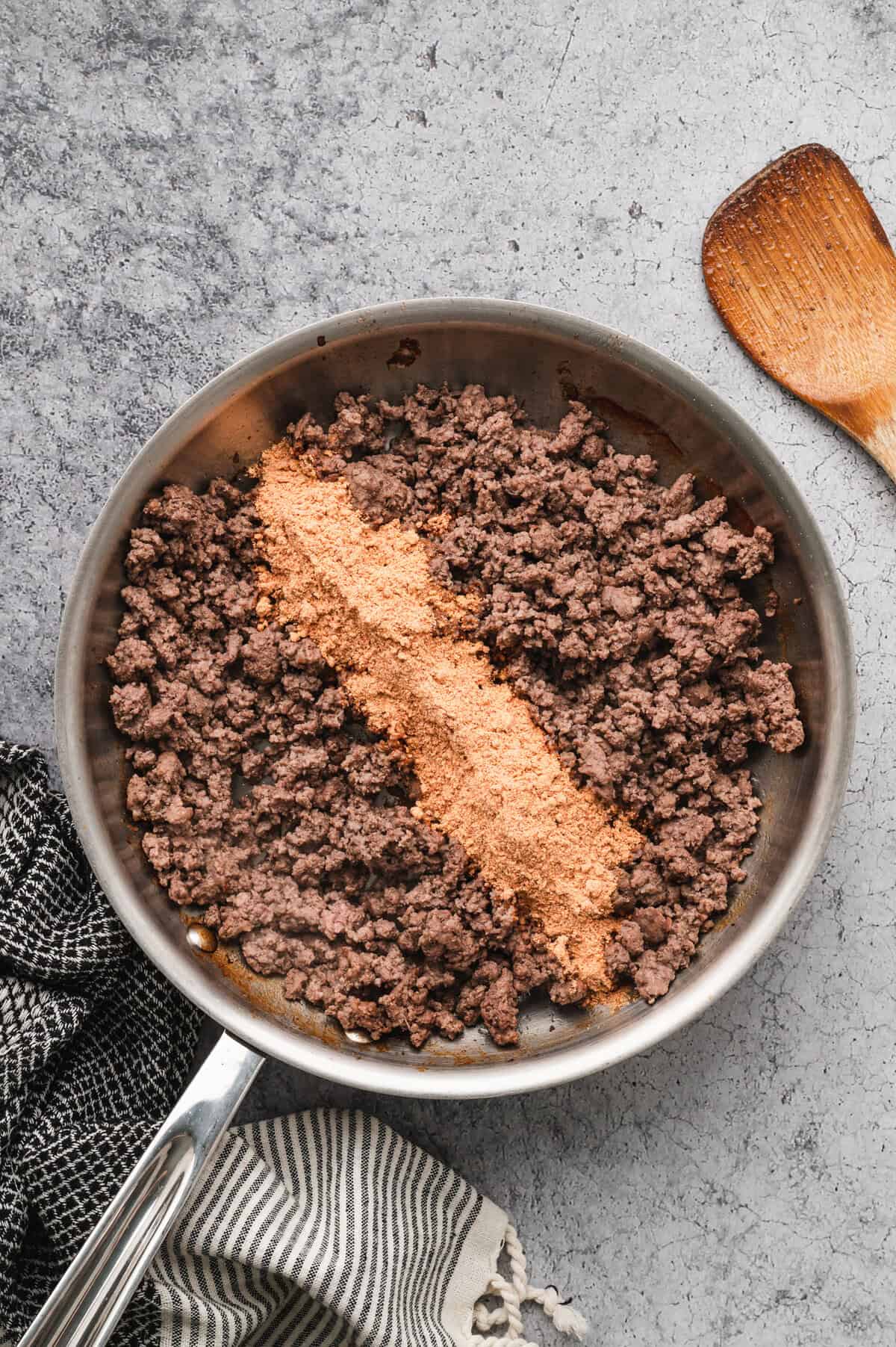 A stainless steel pan filled with cooked ground beef sits on a textured grey surface. A line of orange taco seasoning has been added on top of the beef, ready to be mixed in. A wooden spoon with seasoning residue rests nearby. Black and white striped kitchen towels are draped around the scene.