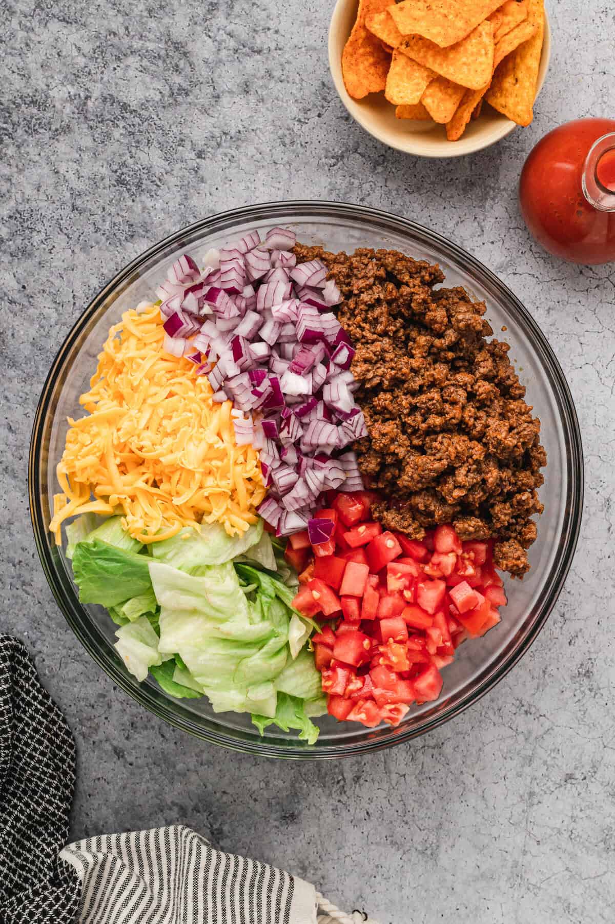 A glass bowl filled with taco salad ingredients sits on a textured grey surface. The bowl contains seasoned ground beef, diced tomatoes, chopped red onions, shredded cheddar cheese, and crisp iceberg lettuce, arranged in separate sections. In the background, a small bowl of Doritos and a bottle of red dressing are visible. Black and white striped kitchen towels are draped around the scene.