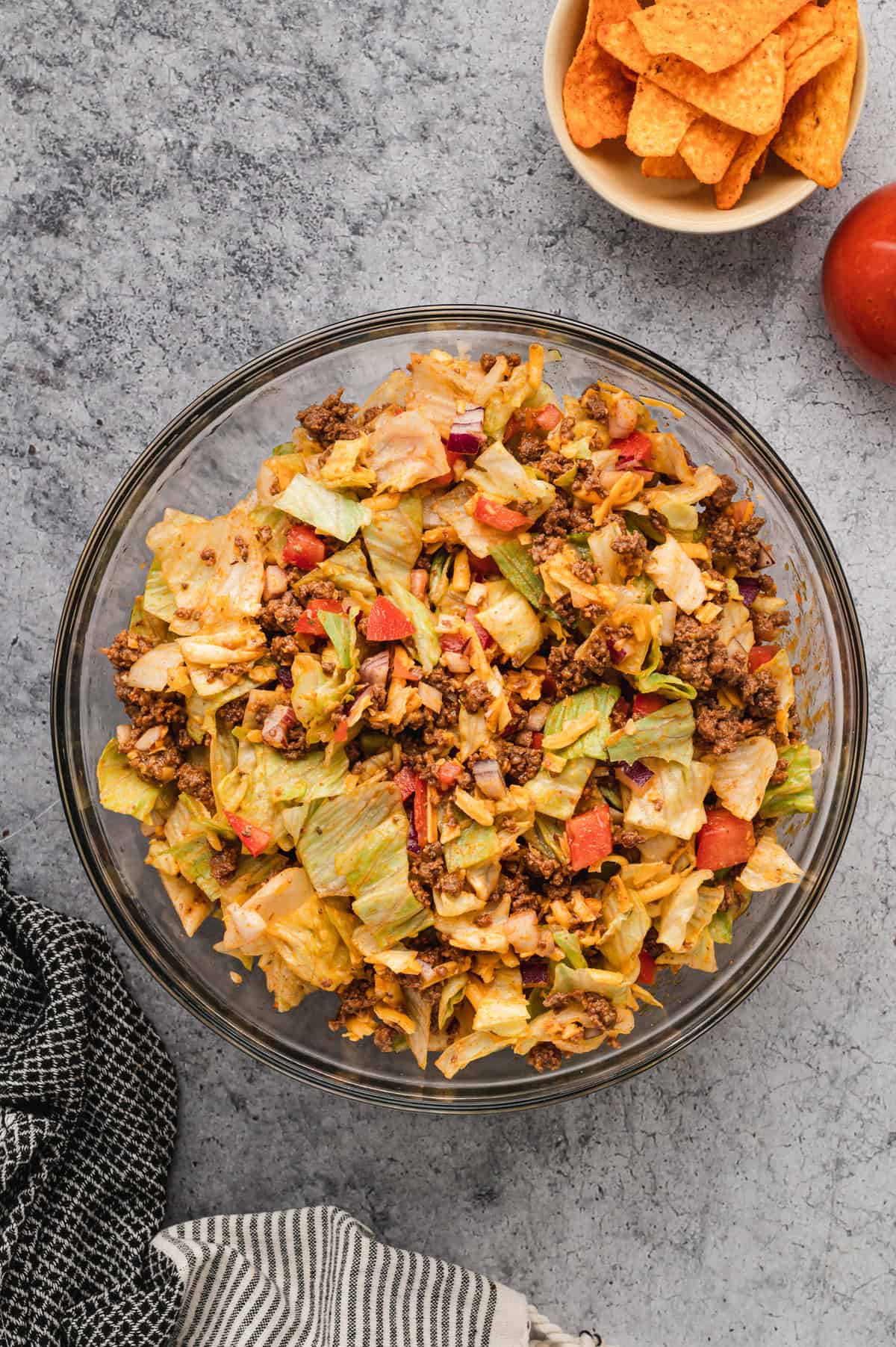 A glass bowl filled with Dorito taco salad sits on a textured grey surface. The salad is fully mixed, combining seasoned ground beef, chopped iceberg lettuce, diced tomatoes, red onions, shredded cheddar cheese, crushed Doritos, and dressing. A small bowl of Doritos and a bottle of red dressing are visible in the background. Black and white striped kitchen towels are draped around the scene.