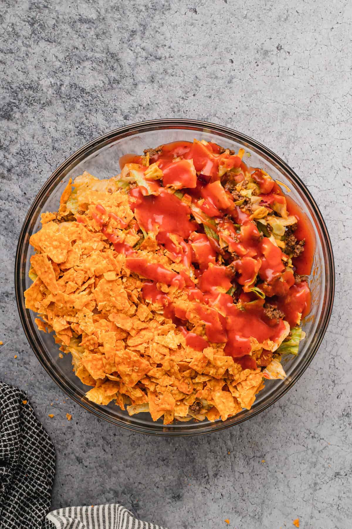 A glass bowl filled with Dorito taco salad sits on a grey textured surface. The salad is topped with crushed Doritos on one side and drizzled with a red dressing on the other. The ingredients, including seasoned ground beef, lettuce, tomatoes, onions, and cheese, are visible beneath the toppings. A black and white striped kitchen towel is placed nearby, with a few stray crumbs scattered around.