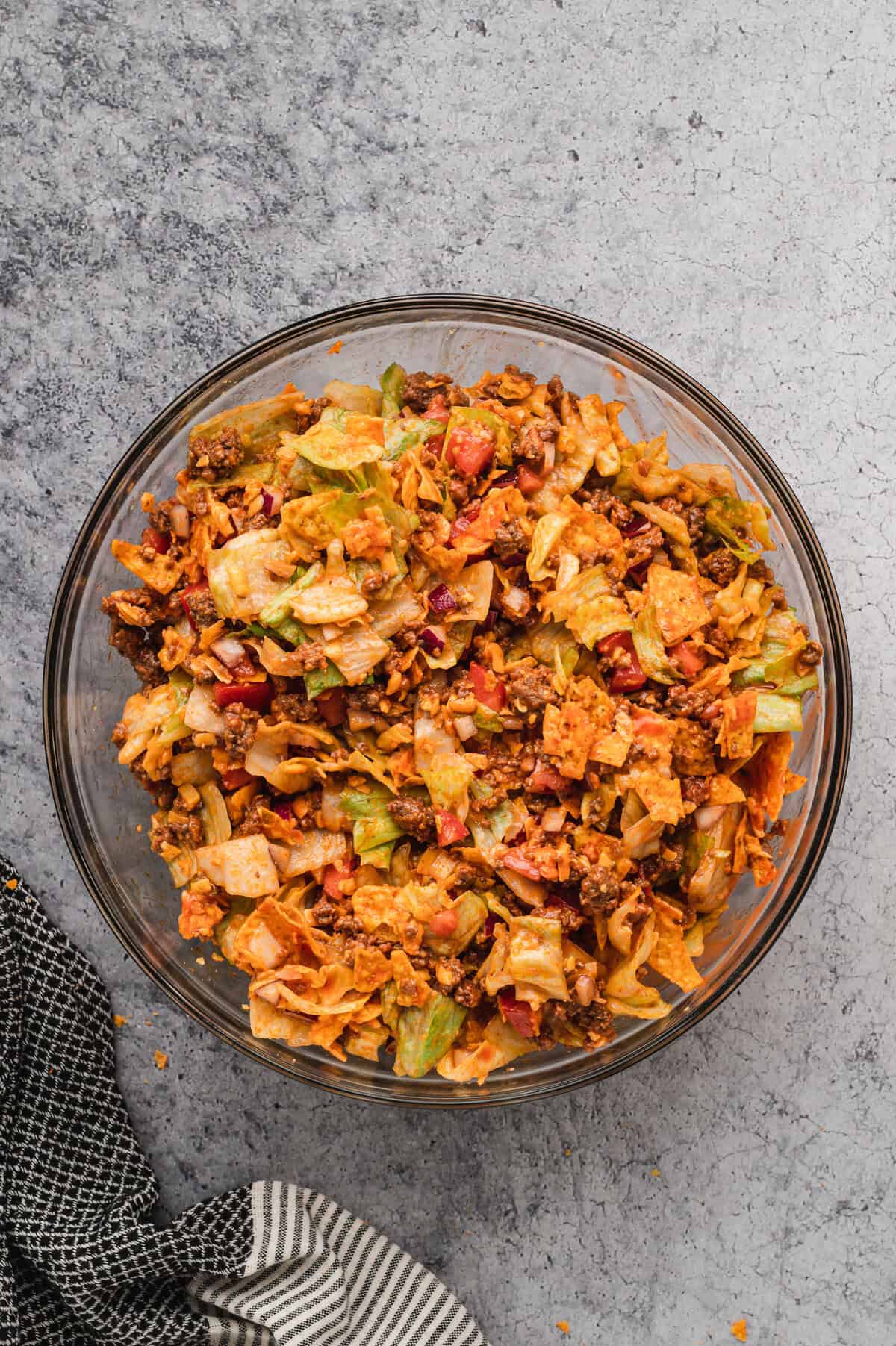 A glass bowl filled with freshly mixed Dorito taco salad sits on a grey textured surface. The salad consists of chopped lettuce, diced tomatoes, red onions, seasoned ground beef, shredded cheese, and crushed Doritos, all coated in a tangy dressing. The ingredients are well combined, with the crunchy chips and vegetables evenly distributed. A black and white striped kitchen towel rests beside the bowl, with a few crumbs scattered around.