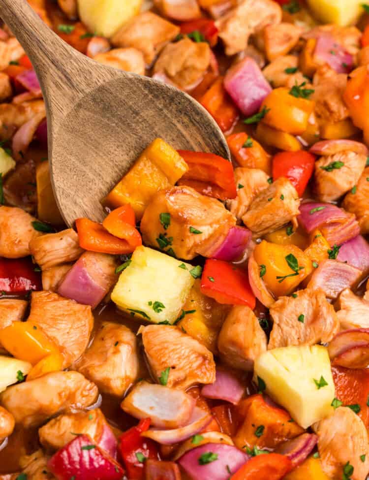 A close-up of Hawaiian sheet pan chicken with bite-sized pieces of chicken, red and yellow bell peppers, red onions, and chunks of pineapple, all coated in a glossy sauce and garnished with chopped parsley. A wooden spoon is scooping up a portion of the dish.