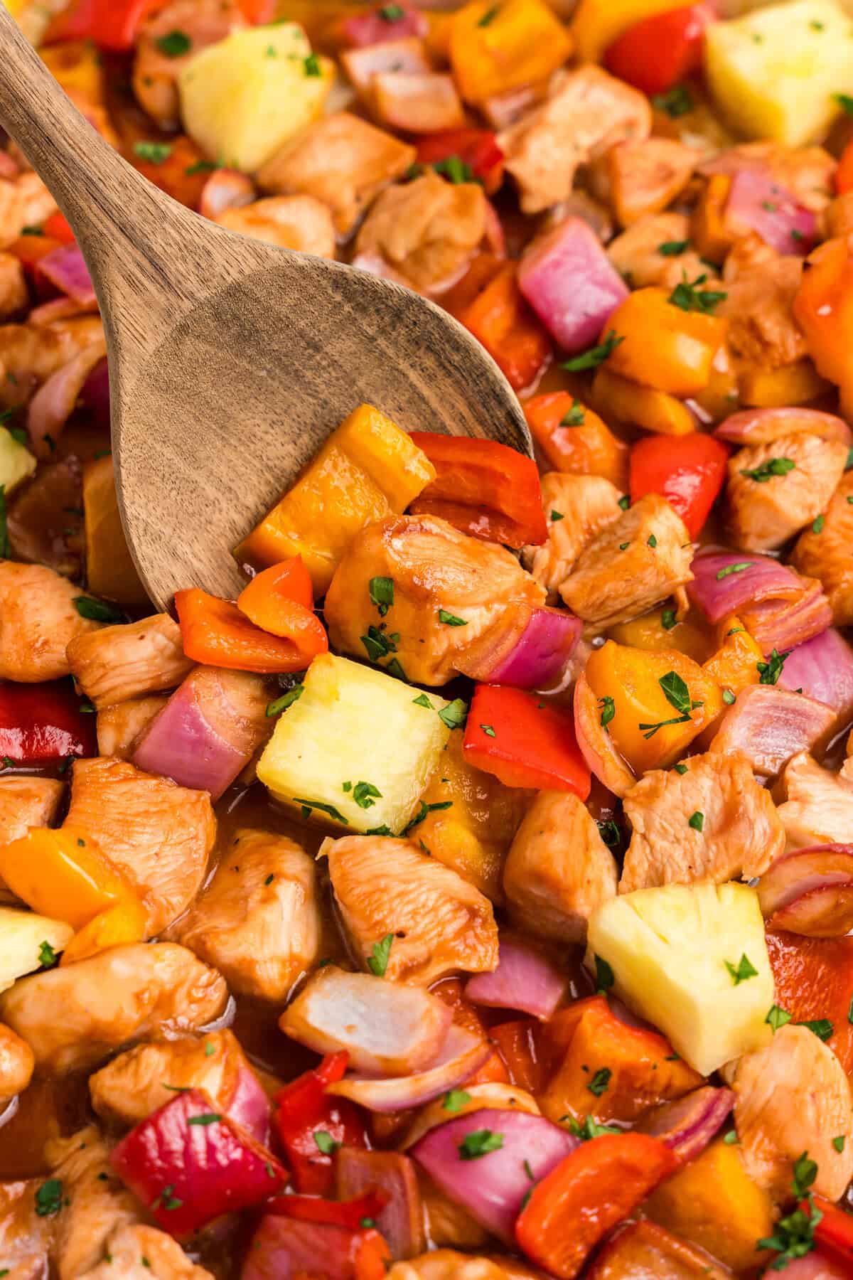 A close-up of Hawaiian sheet pan chicken with bite-sized pieces of chicken, red and yellow bell peppers, red onions, and chunks of pineapple, all coated in a glossy sauce and garnished with chopped parsley. A wooden spoon is scooping up a portion of the dish.