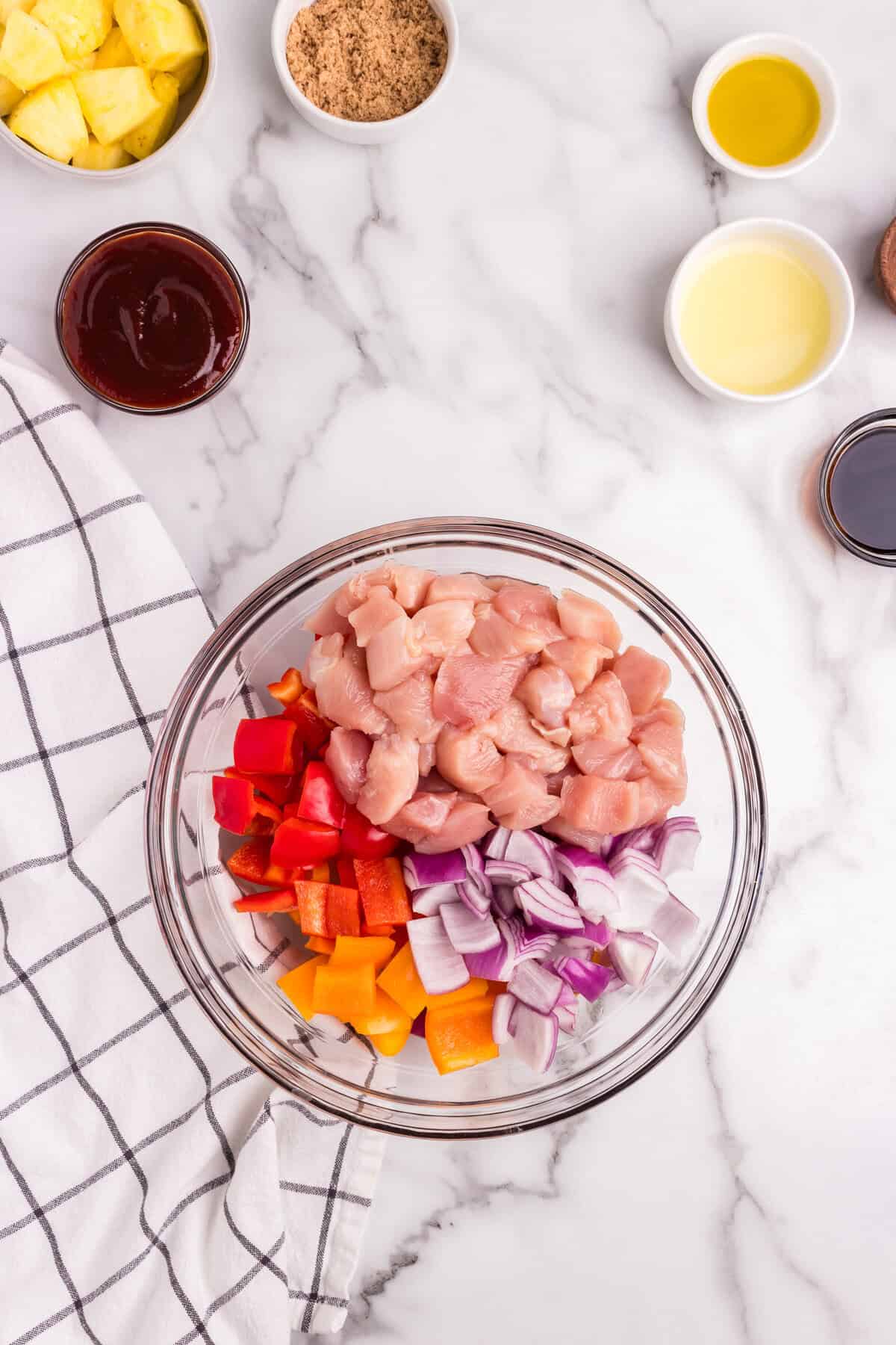 A glass mixing bowl filled with diced raw chicken, chopped red and yellow bell peppers, and diced red onions on a marble countertop. Surrounding the bowl are small bowls containing brown sugar, barbecue sauce, pineapple chunks, olive oil, and another liquid ingredient.