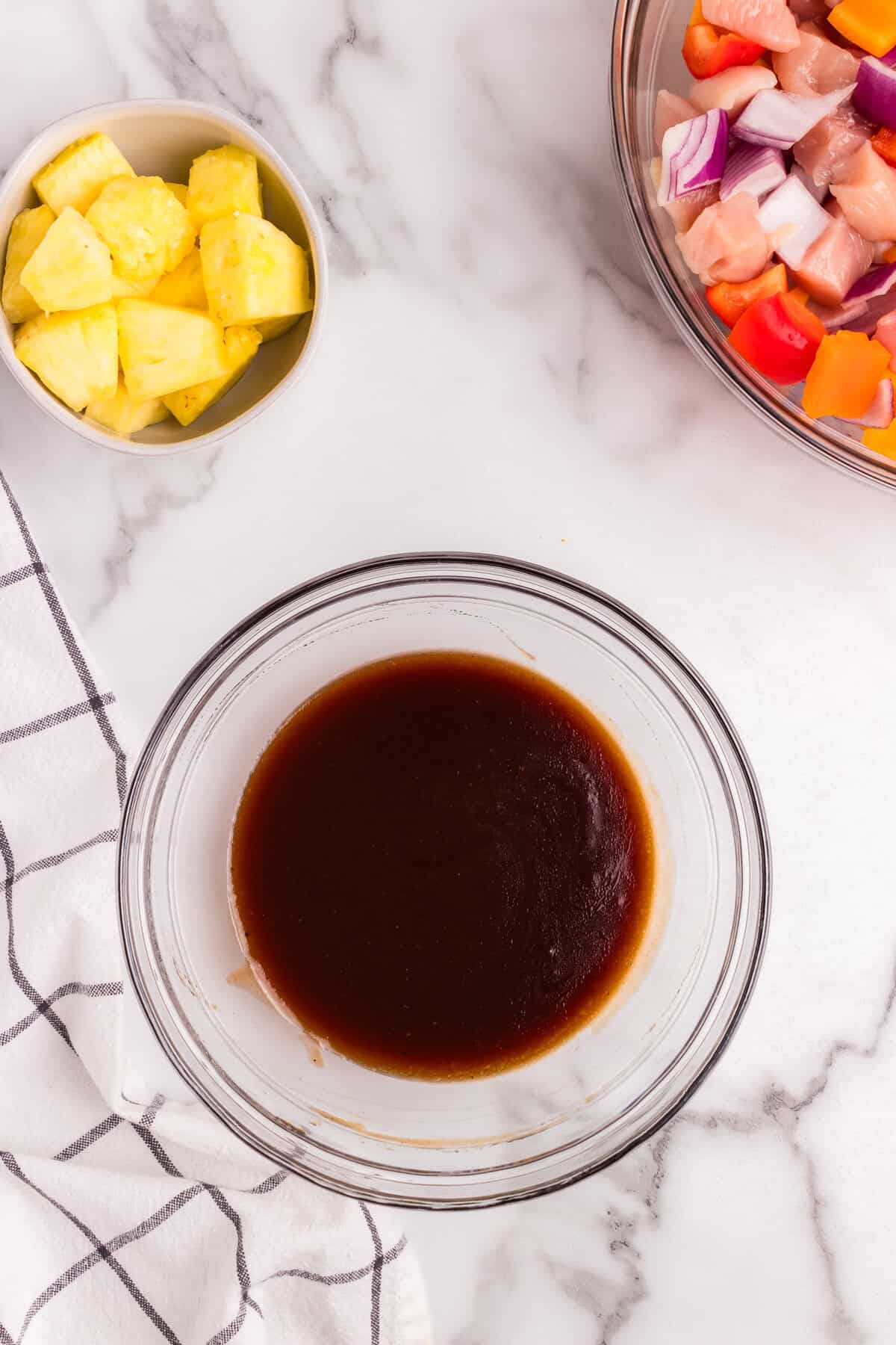 A separate glass bowl containing a dark barbecue sauce mixture on a marble countertop. A bowl of fresh pineapple chunks is placed nearby.