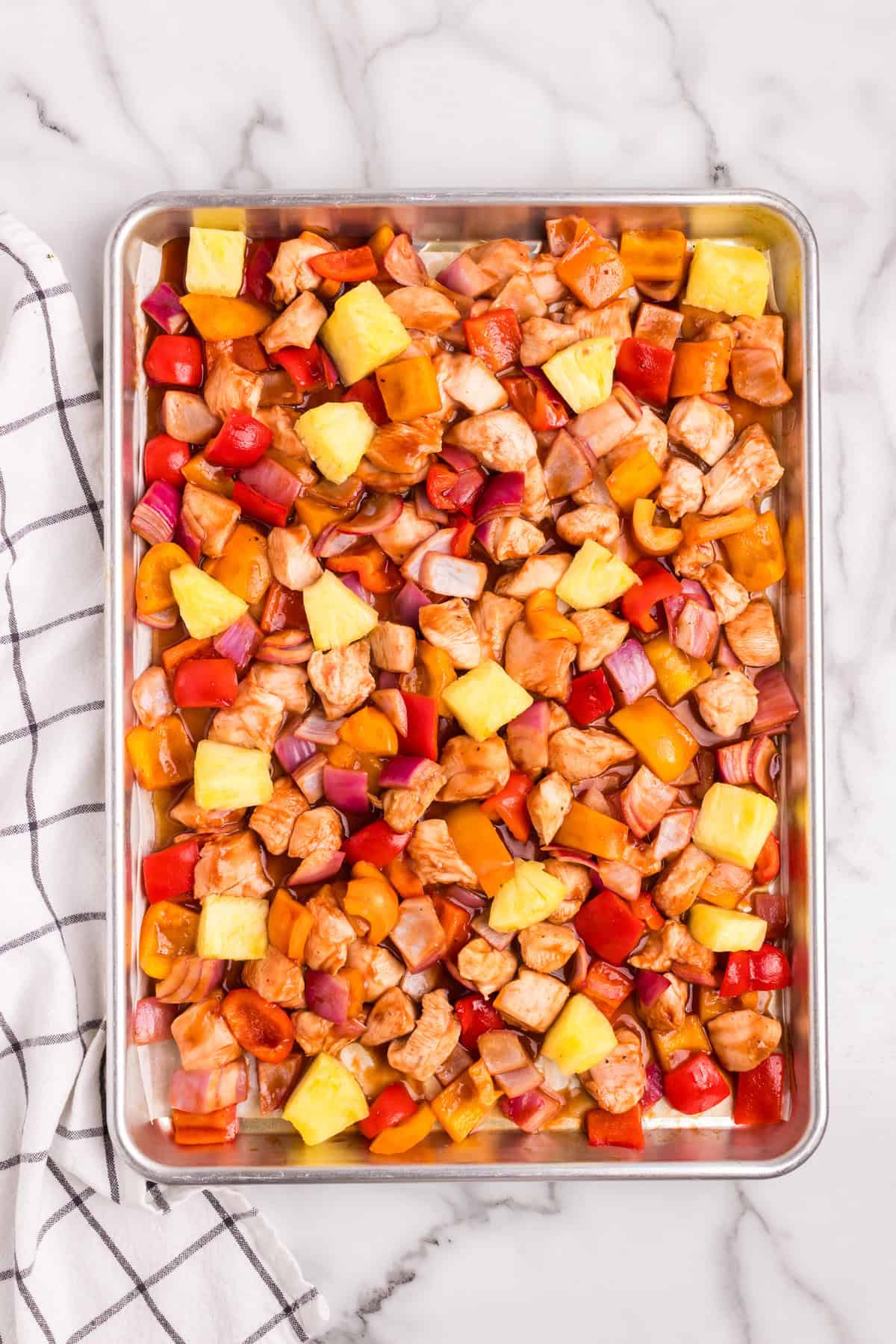 A sheet pan filled with marinated diced chicken, bell peppers, red onions, and pineapple chunks, evenly spread out and ready for baking.