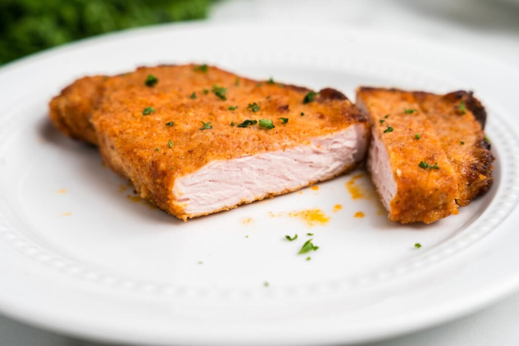 A close-up of a golden-brown, breaded pork chop sliced in half on a white plate, garnished with chopped parsley.