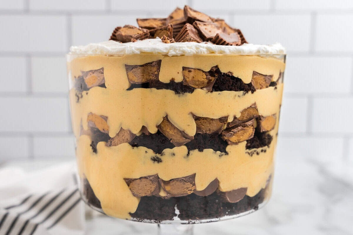  A close-up of a fully assembled Reese’s Peanut Butter Trifle in a clear glass dish, showcasing layers of brownies, peanut butter filling, and chopped Reese’s cups topped with whipped cream.