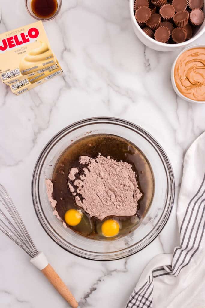 A bowl with brownie mix, eggs, and oil ready to be mixed, surrounded by ingredients like vanilla pudding mix, peanut butter, Reese’s cups, and vanilla extract.