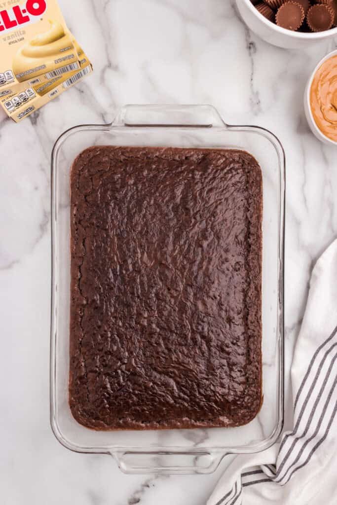 A pan of freshly baked brownies cooling on a marble countertop, with pudding mix, peanut butter, and Reese’s cups visible nearby.