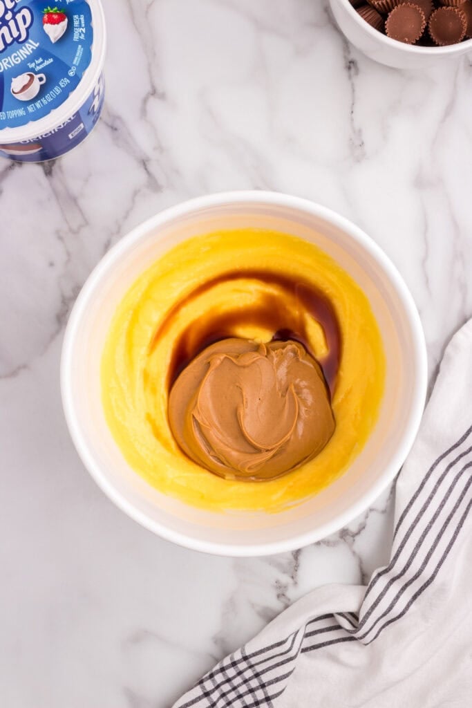 A mixing bowl containing vanilla pudding, peanut butter, and vanilla extract before being blended, with a container of whipped topping in the background.