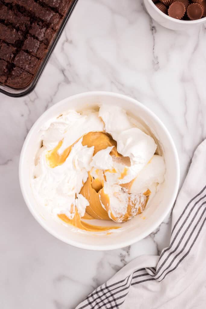 A bowl showing whipped topping being folded into the peanut butter mixture, with brownies and Reese’s cups visible in the background.