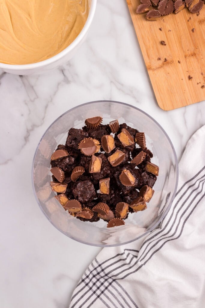 A trifle dish being filled with a layer of chopped brownies and Reese’s peanut butter cups, with peanut butter mixture and cutting board nearby.