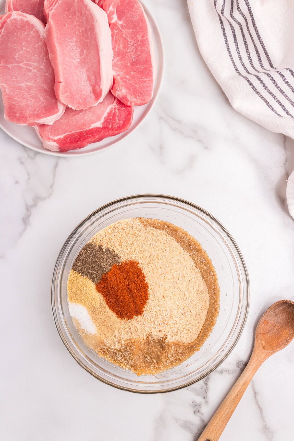 A plate of raw boneless pork chops next to a glass bowl containing a mixture of breadcrumbs and seasonings.