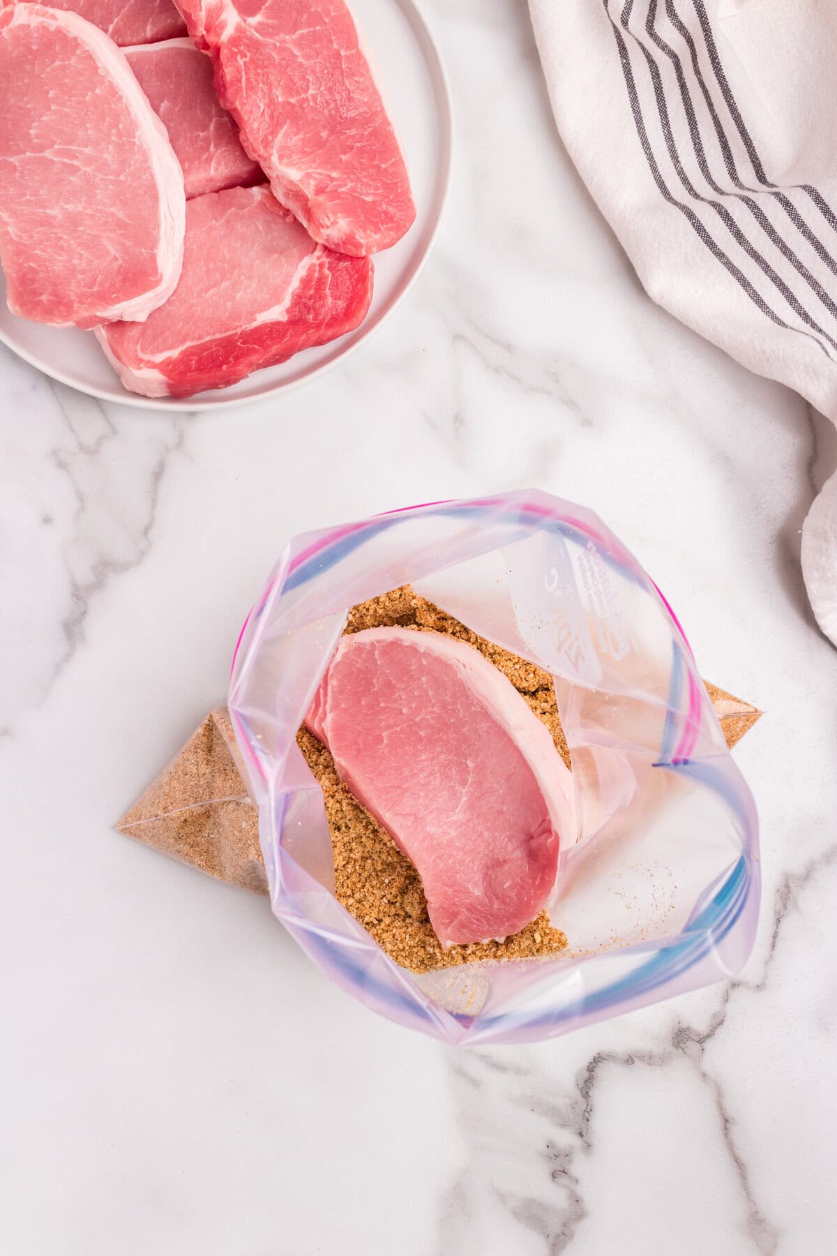 A raw pork chop being placed into a plastic bag containing the breadcrumb mixture for coating.