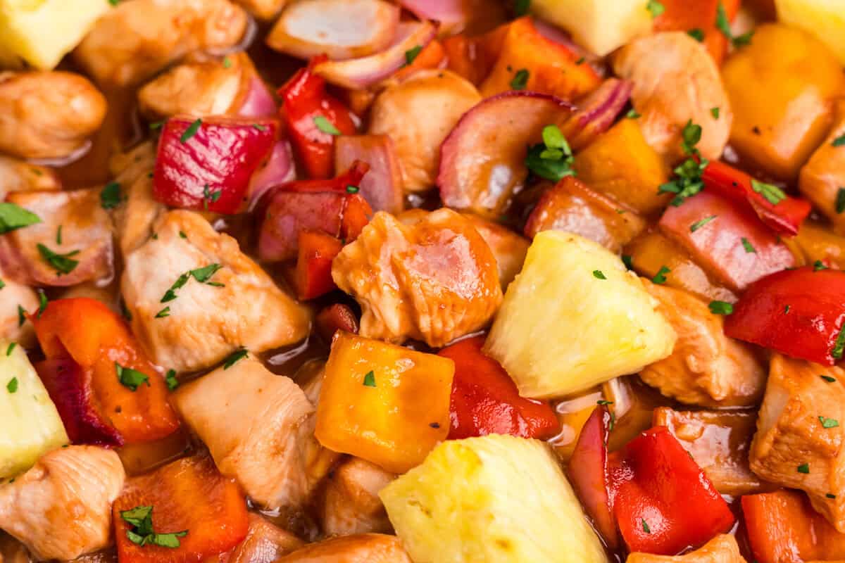 A close-up shot of the finished Hawaiian sheet pan chicken, featuring glistening pieces of chicken, red and orange bell peppers, red onion, and pineapple chunks, garnished with freshly chopped parsley.