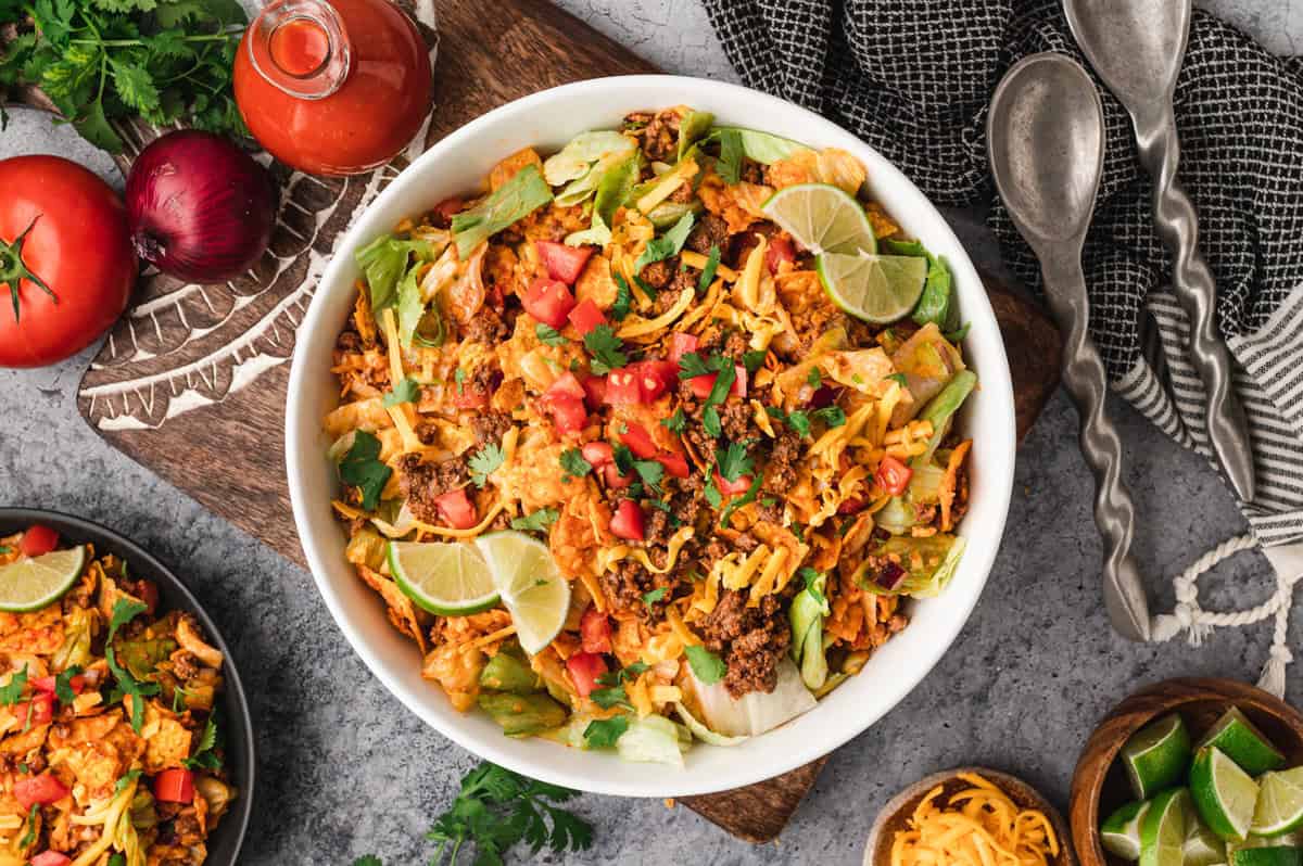 A large white bowl filled with Dorito taco salad, garnished with fresh cilantro, diced tomatoes, shredded cheddar cheese, and lime wedges. The salad contains crunchy Doritos, seasoned ground beef, and crisp lettuce. The bowl is placed on a rustic wooden board, surrounded by fresh ingredients like tomatoes, red onions, a bottle of taco sauce, and lime wedges. A black plate with a serving of the salad is also visible, along with decorative utensils and a textured napkin, creating a vibrant and inviting presentation.
