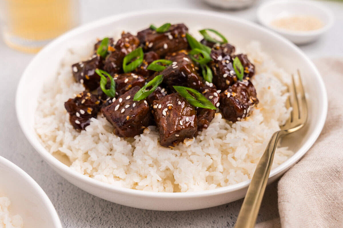 A bowl of white rice topped with the finished teriyaki beef, garnished with sesame seeds and chopped green onions, served with a gold fork.