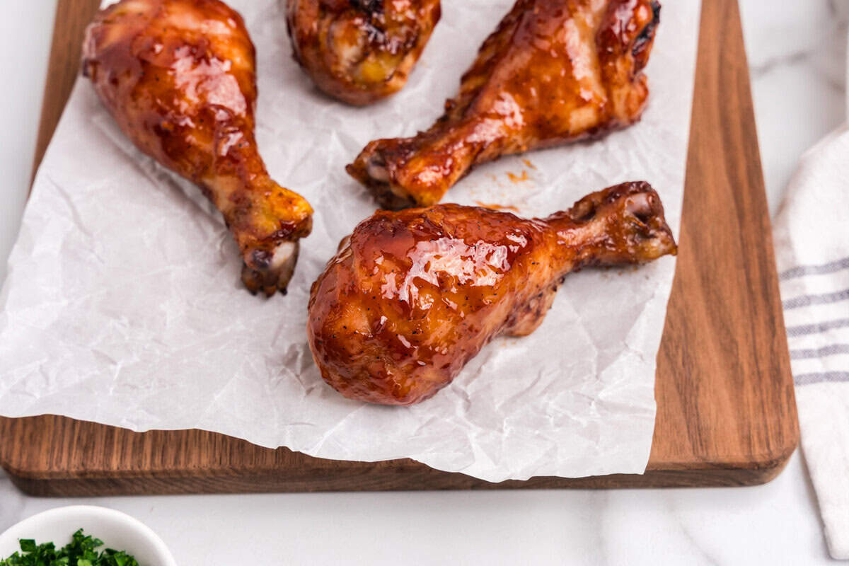 Close-up of four glazed BBQ chicken drumsticks resting on crinkled parchment paper atop a wooden serving board. The chicken is richly coated in a glossy barbecue sauce, showing a caramelized, sticky texture. A small bowl of chopped herbs is partially visible in the corner.