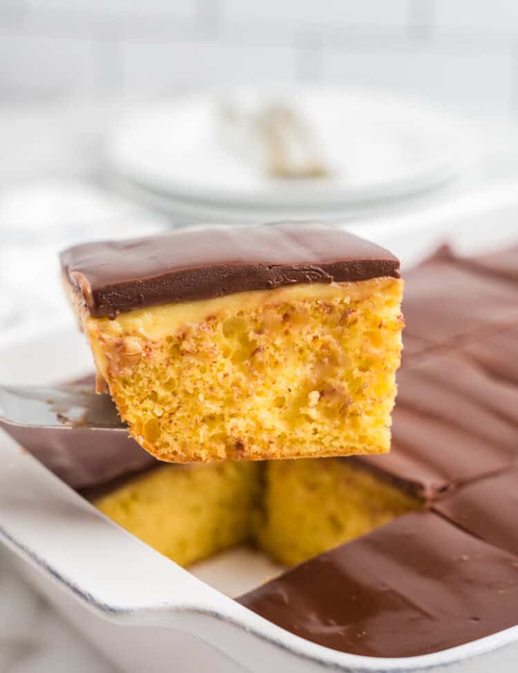 A single piece of Boston Cream Poke Cake held above the dish, showing the layers of moist yellow cake, pudding filling in the poked holes, and glossy chocolate topping.