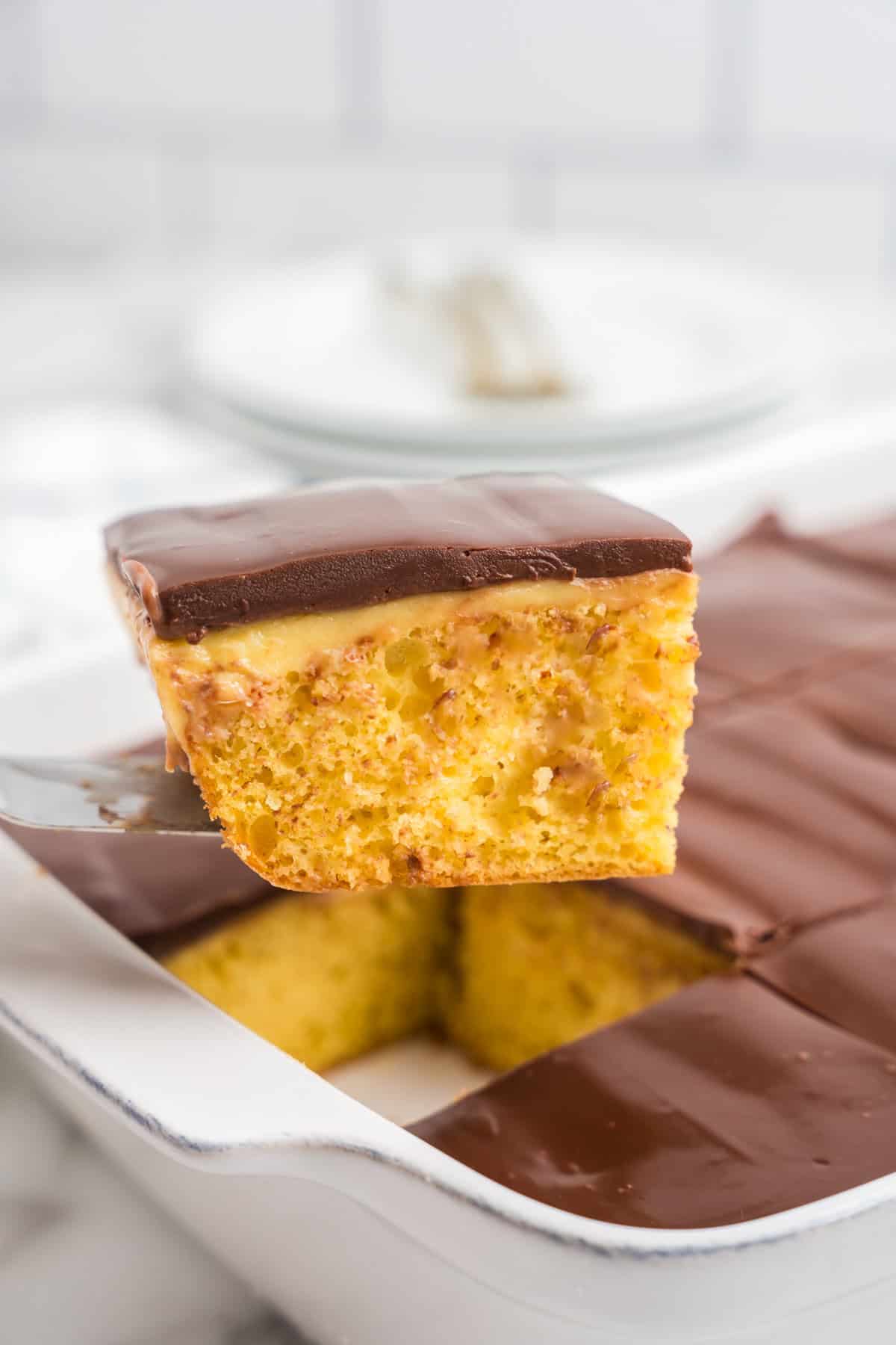 A single piece of Boston Cream Poke Cake held above the dish, showing the layers of moist yellow cake, pudding filling in the poked holes, and glossy chocolate topping.