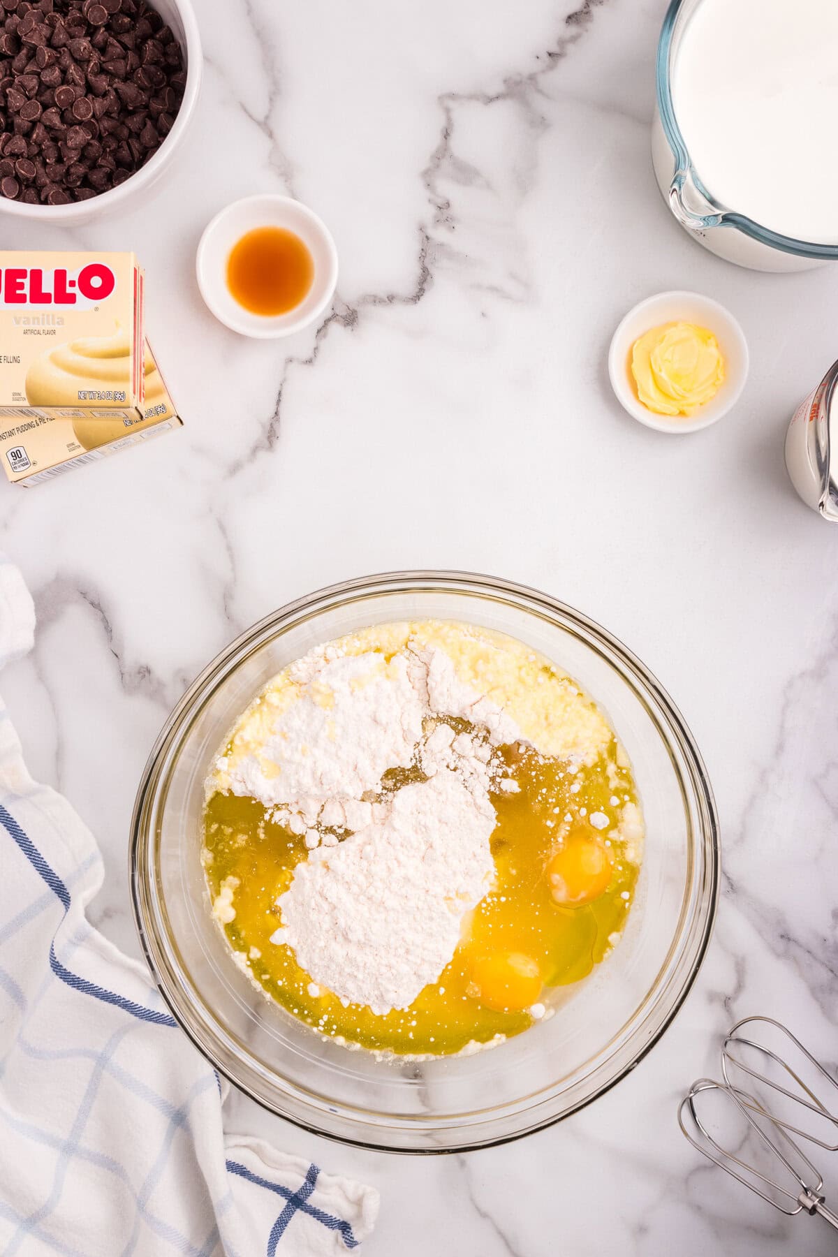 A mixing bowl filled with cake mix, eggs, and other ingredients ready to be mixed. Surrounding the bowl are chocolate chips, vanilla extract, butter, and vanilla pudding mix on a marble countertop.