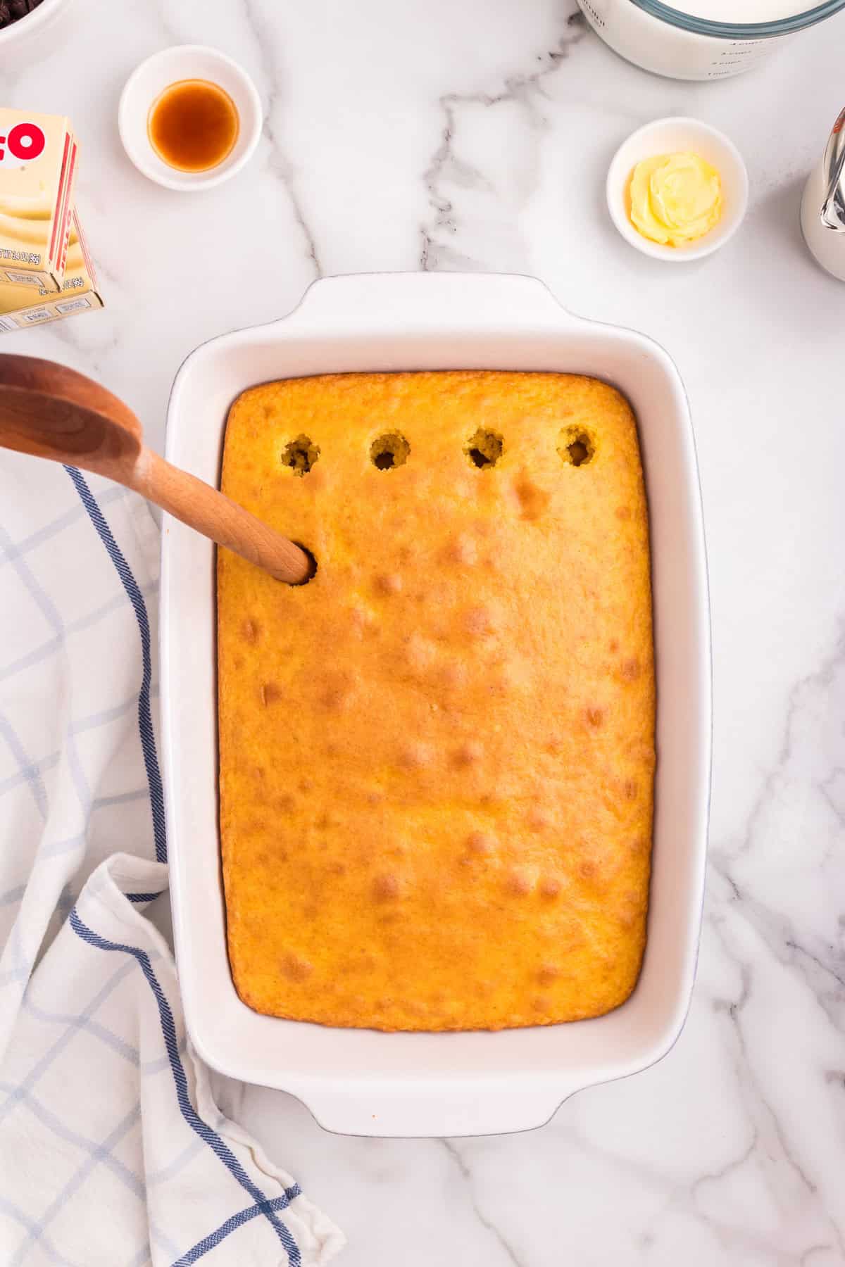 The baked yellow cake in the dish, with a wooden spoon poking large holes into the top surface.