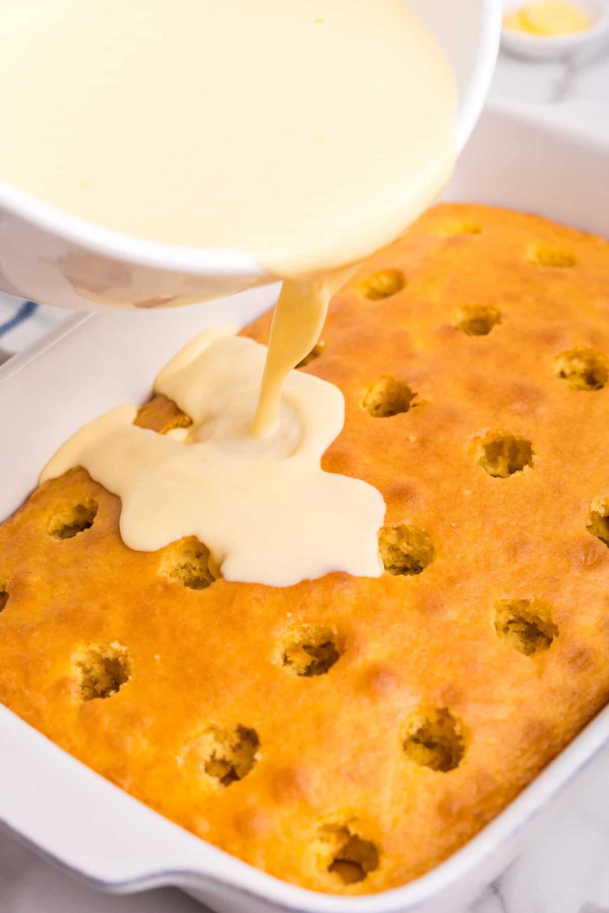 Vanilla pudding being poured from a bowl over a freshly baked cake with evenly spaced holes, filling the holes to create a Boston Cream Poke Cake.