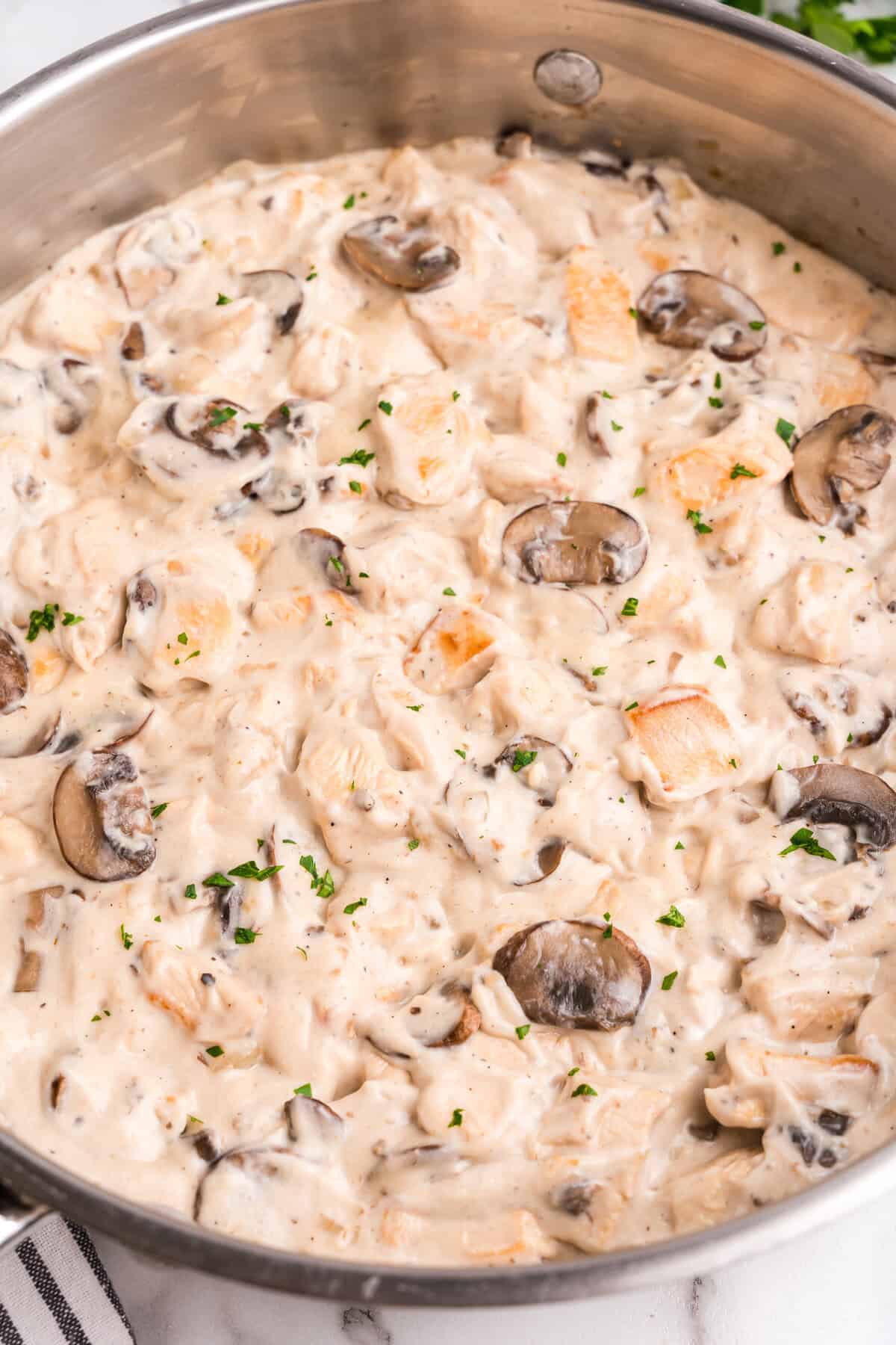 A creamy chicken stroganoff dish in a stainless steel skillet, featuring chunks of cooked chicken, sliced mushrooms, and a rich white sauce, garnished with chopped parsley.