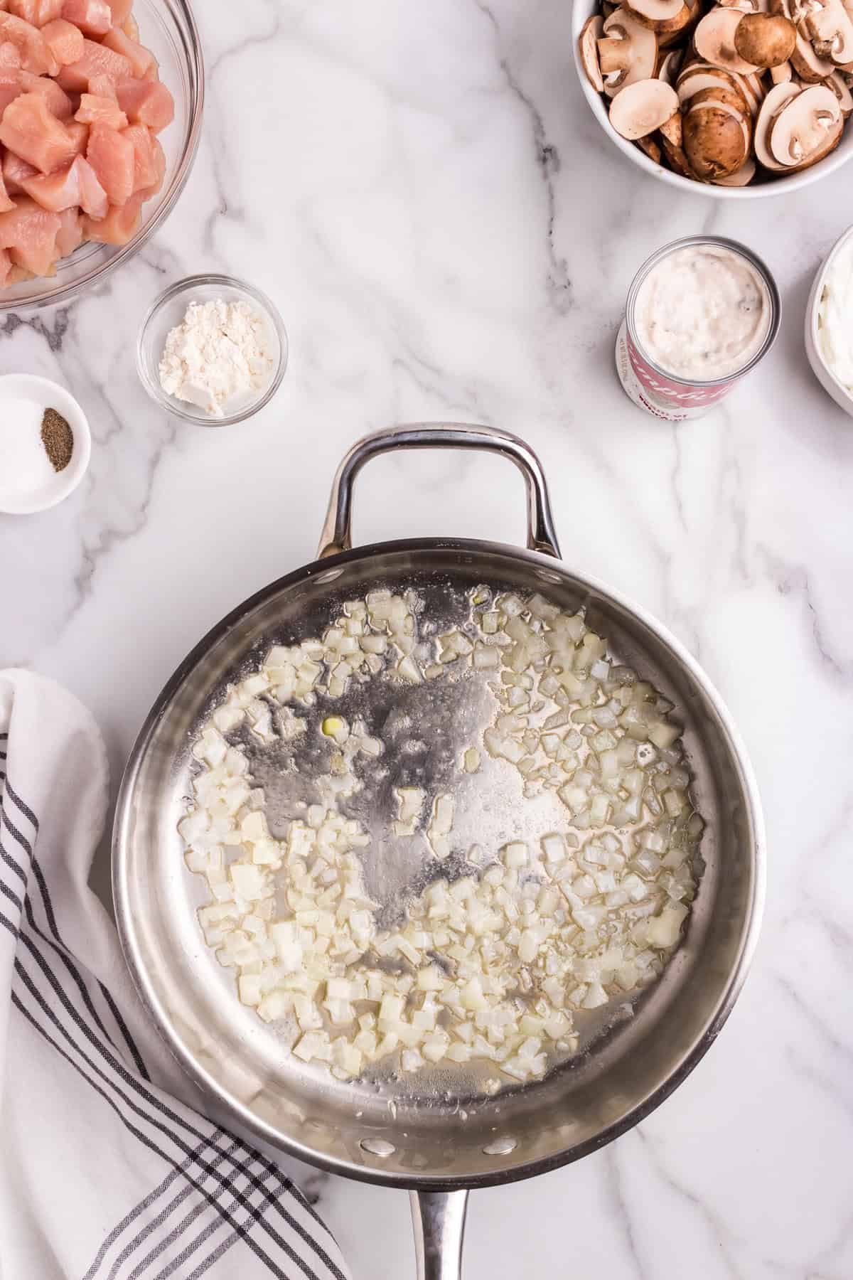 Diced onions sautéing in a stainless steel skillet with oil, surrounded by raw chicken, sliced mushrooms, cream of mushroom soup, sour cream, flour, salt, and pepper on a marble countertop.