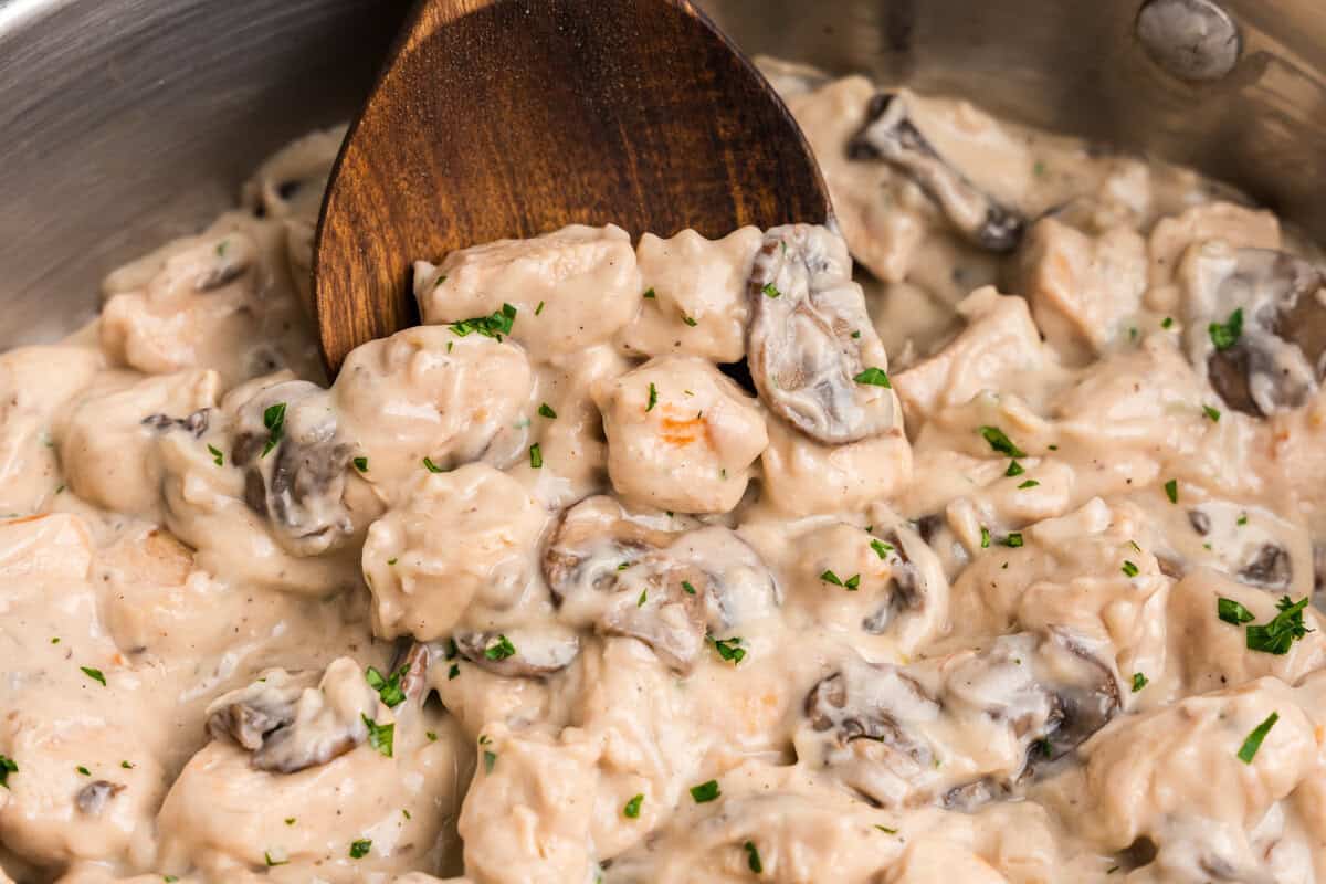 Close-up of creamy chicken stroganoff being stirred with a wooden spoon in a stainless steel pan. The dish features tender chunks of chicken, sliced mushrooms, and a rich, creamy sauce, garnished with chopped parsley.
