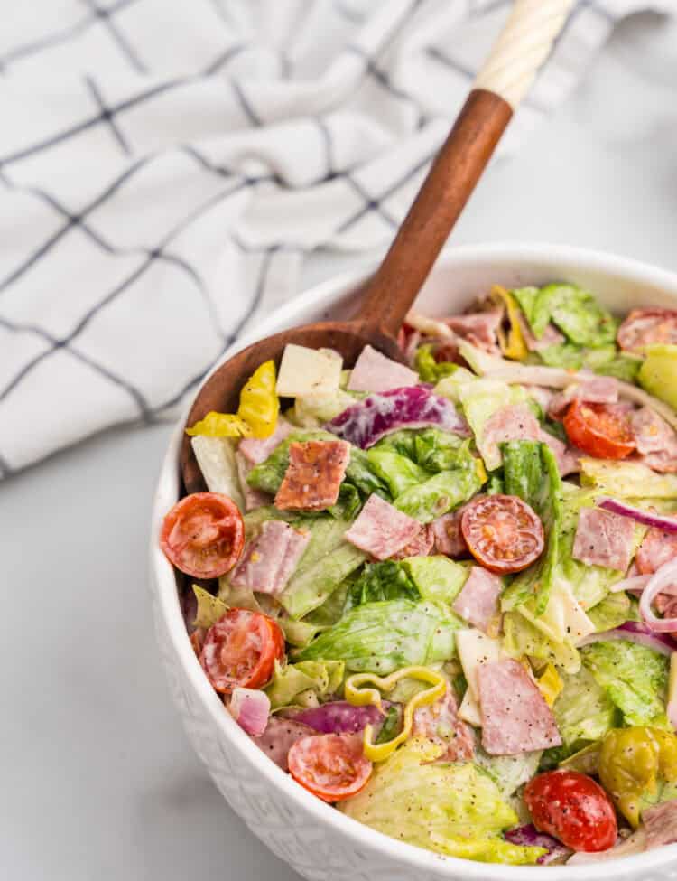 A white textured bowl filled with grinder salad and a wooden serving spoon, with a dish towel in the background for a rustic kitchen aesthetic.