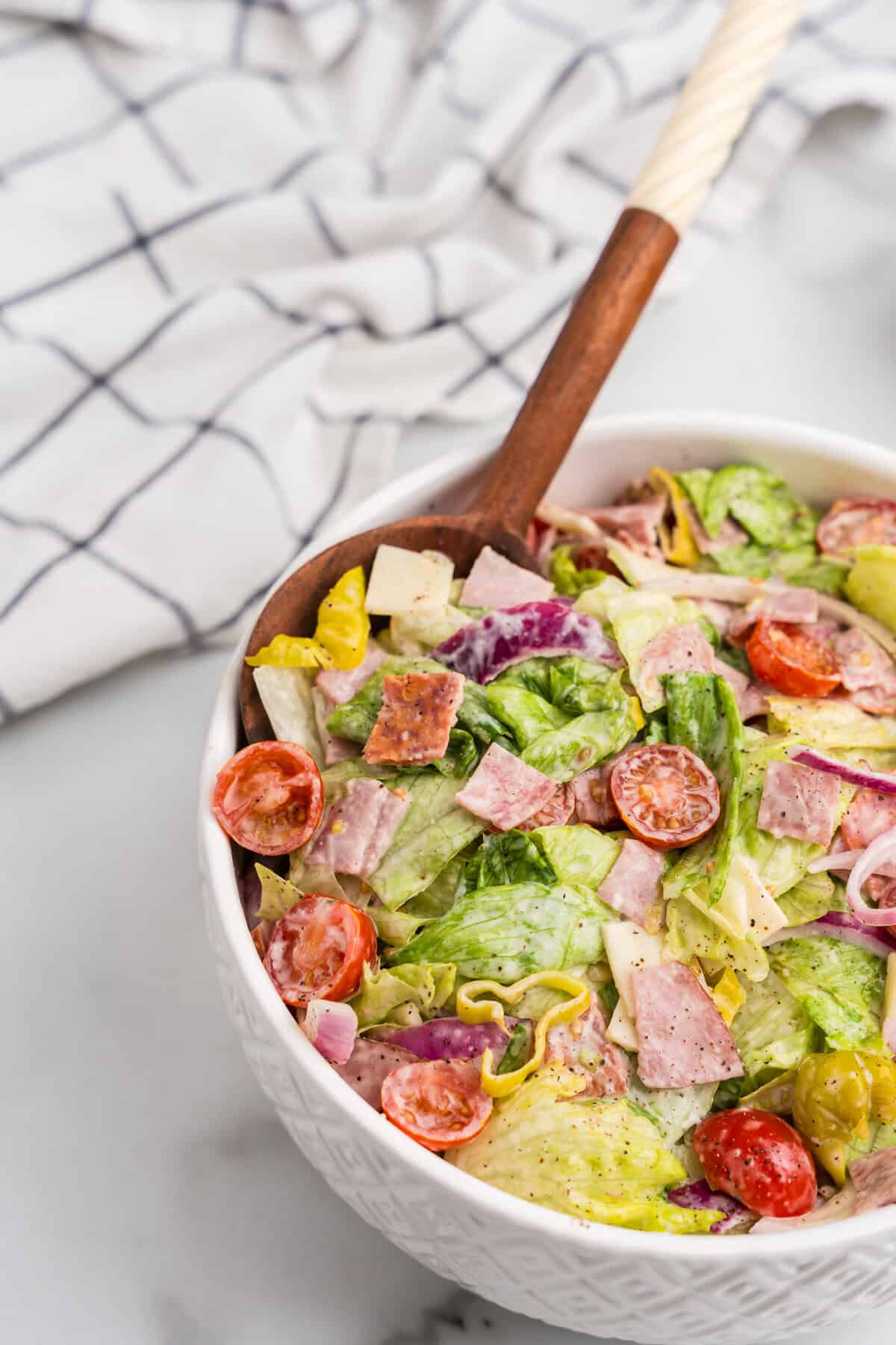 A white textured bowl filled with grinder salad and a wooden serving spoon, with a dish towel in the background for a rustic kitchen aesthetic.