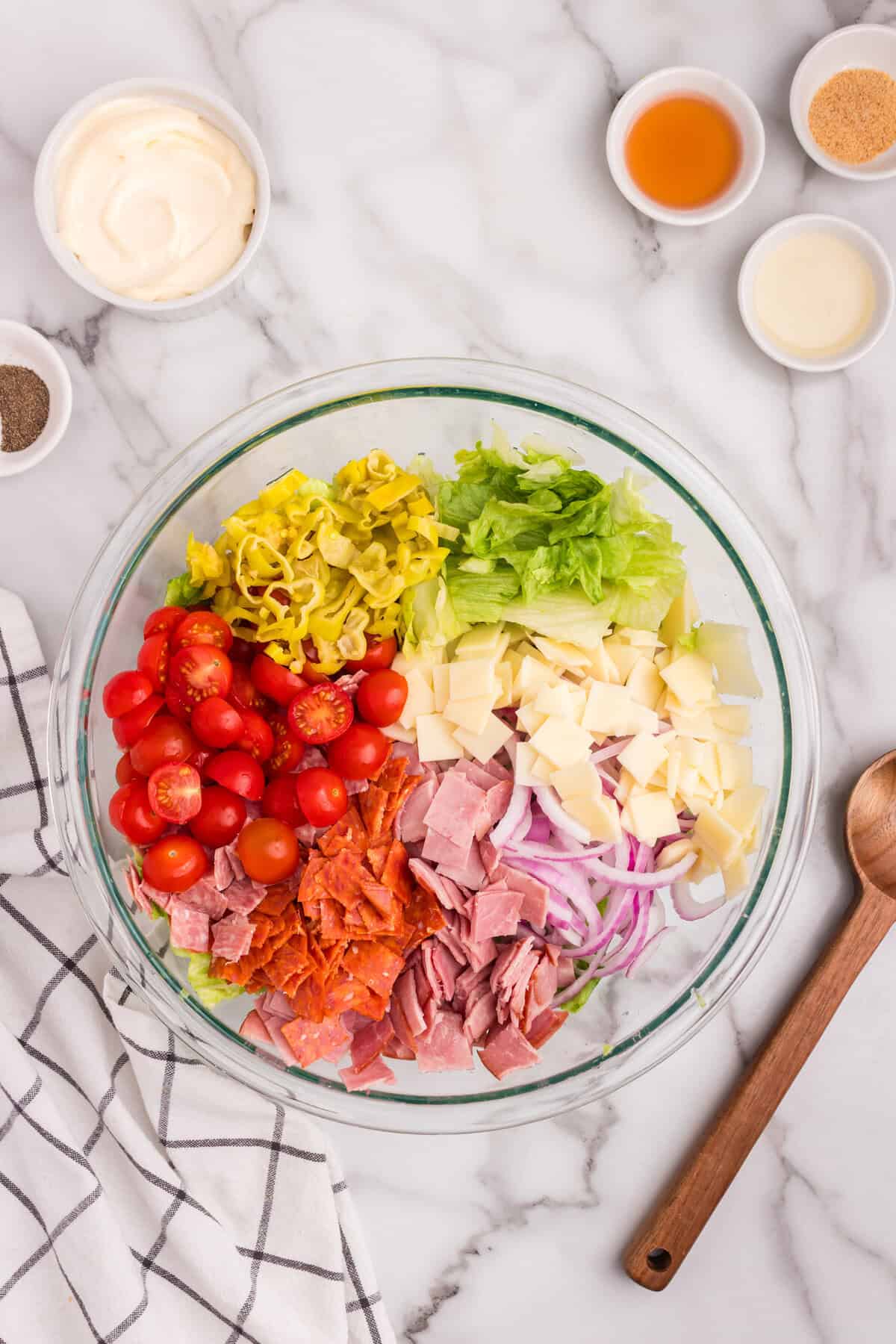 A large glass bowl with neatly arranged salad ingredients including shredded lettuce, halved cherry tomatoes, banana peppers, sliced red onions, cubed provolone cheese, chopped ham, salami, and pepperoni.