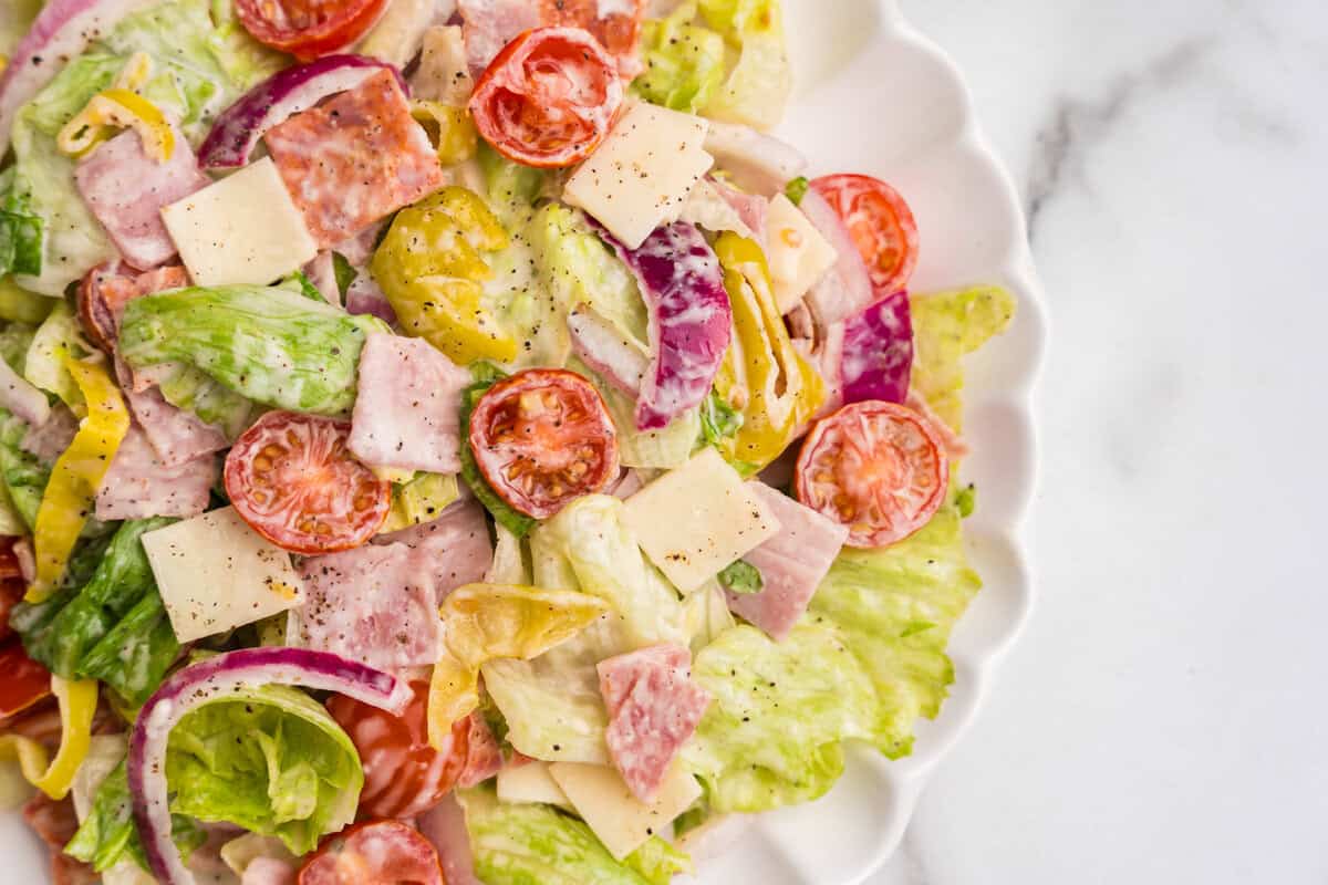 A close-up of a serving plate filled with grinder salad, showcasing crisp lettuce, juicy cherry tomatoes, pickled banana peppers, red onions, deli meats, and cubes of cheese coated in creamy dressing.