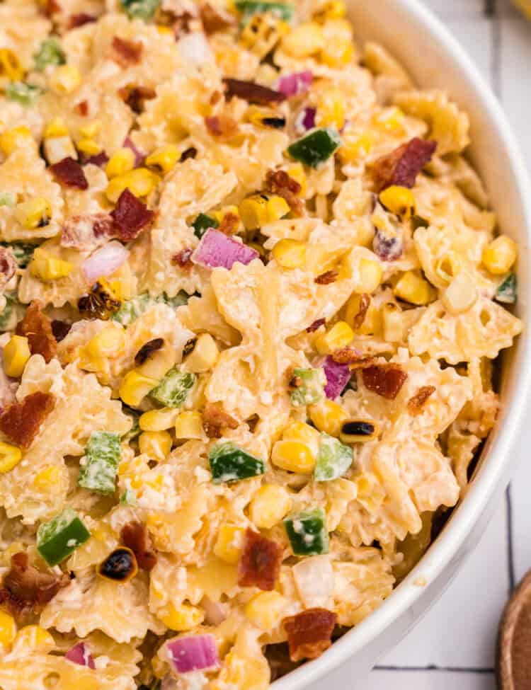 A close-up view highlighting textures and colors: grilled corn, creamy pasta, crispy bacon, red onion, and green pepper.
