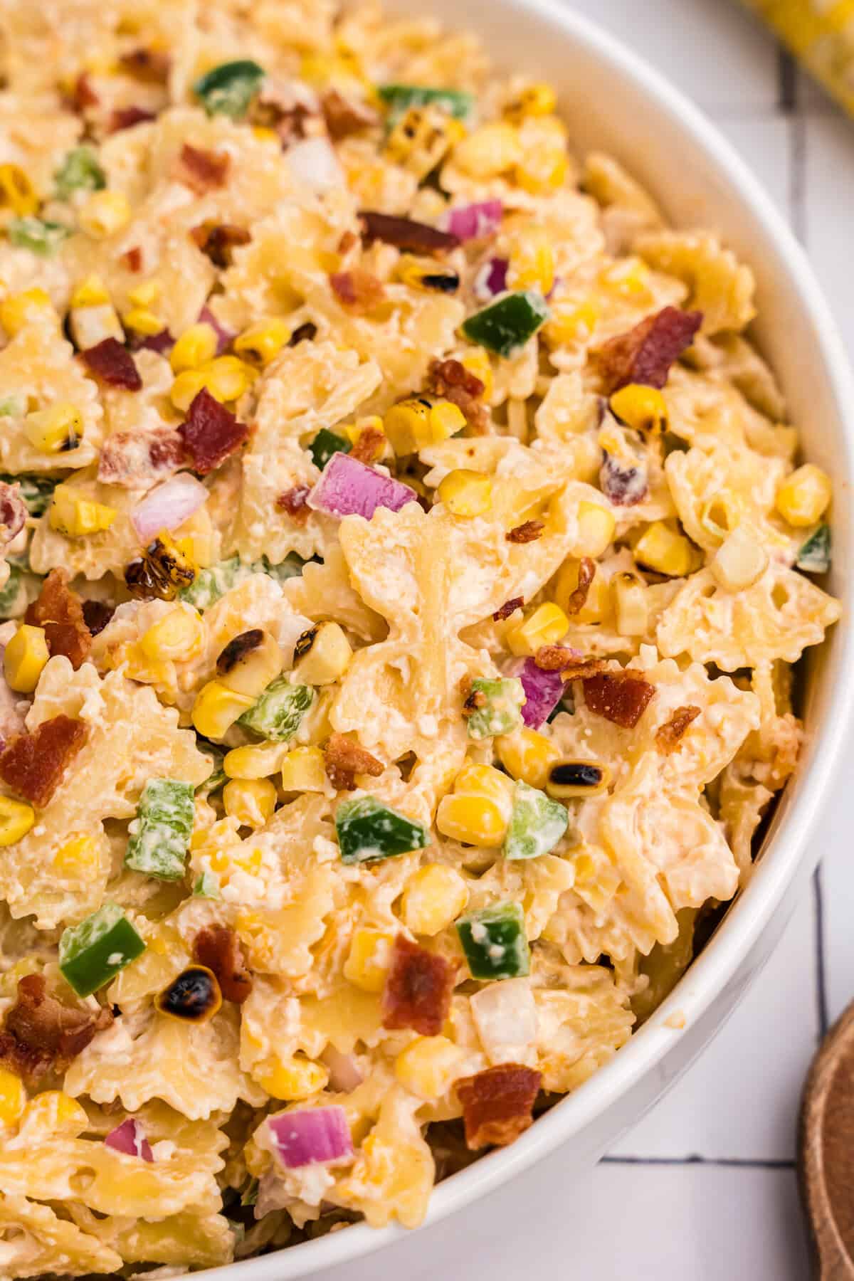 A close-up view highlighting textures and colors: grilled corn, creamy pasta, crispy bacon, red onion, and green pepper.