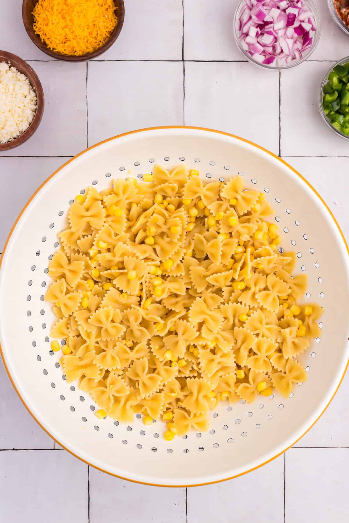 A colander filled with cooked bowtie pasta and yellow corn, set on a tiled surface, ready for mixing.