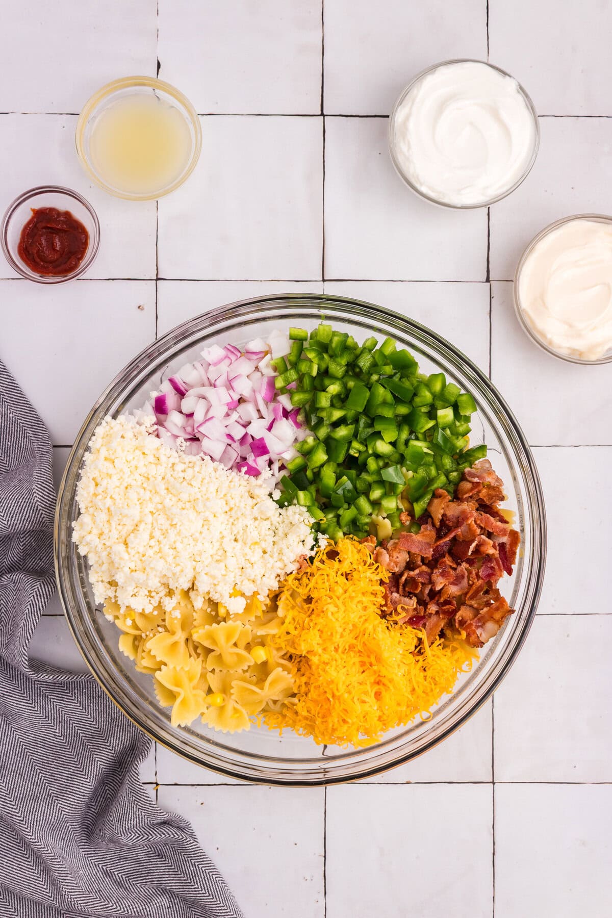 A clear glass bowl containing separated ingredients: chopped red onions, green bell peppers, crispy bacon, shredded cheddar, crumbled cheese, pasta, and corn, surrounded by small bowls of sour cream, mayo, chipotle, and lime juice.