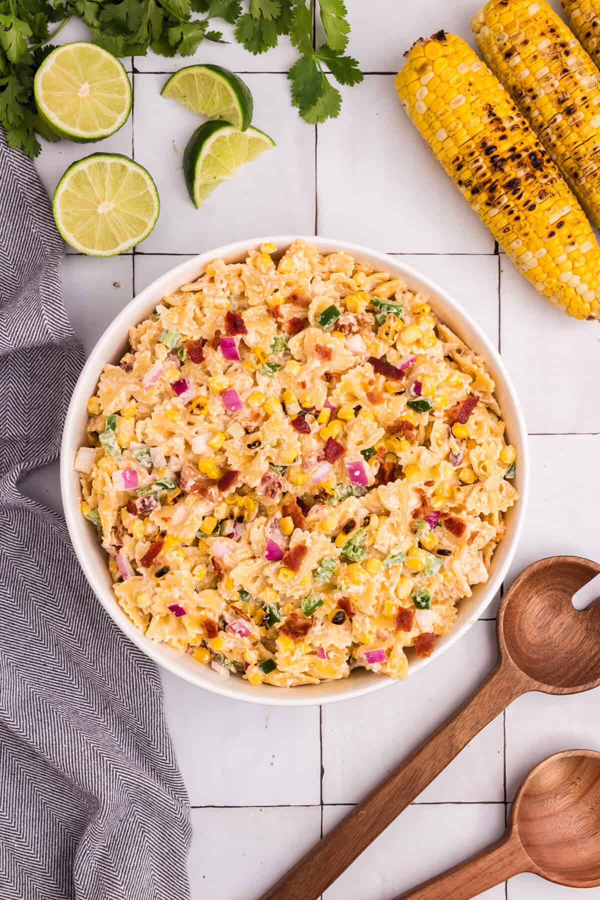 The finished pasta salad served in a white bowl, garnished with cilantro and bacon bits, surrounded by lime wedges and grilled corn.