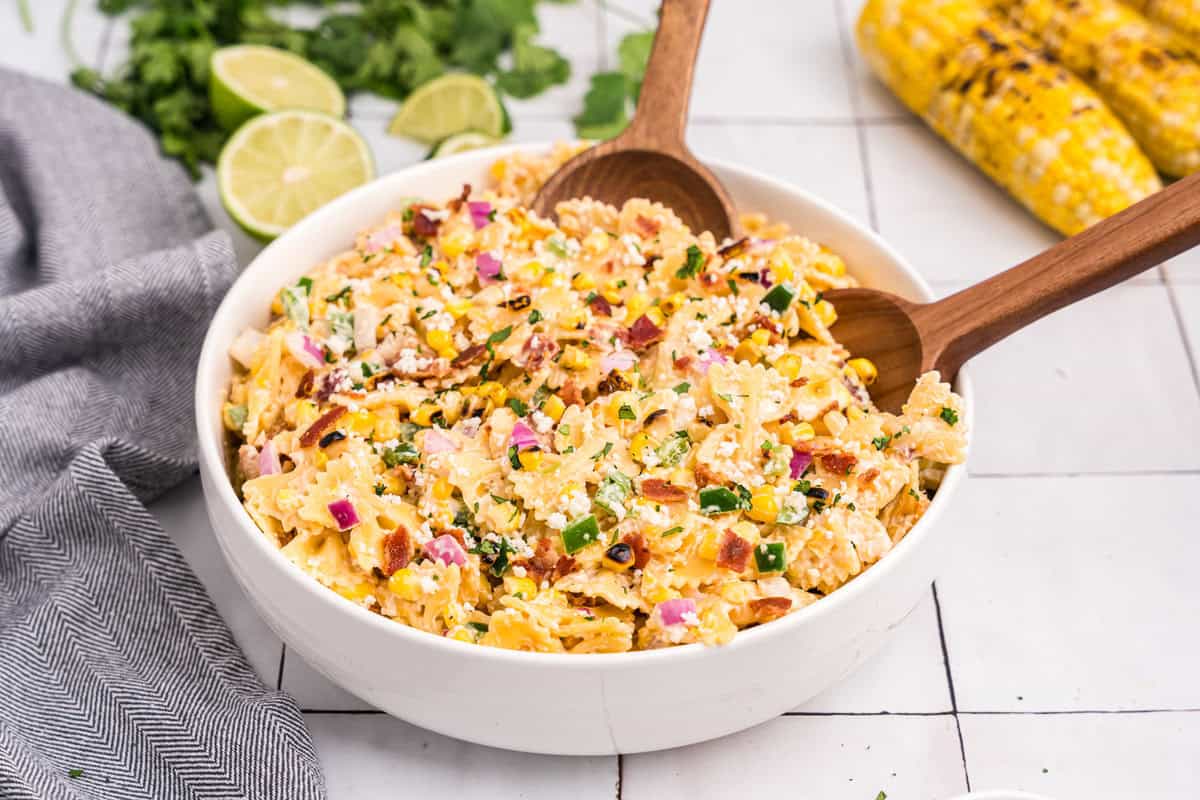 A large serving bowl filled with the completed salad, wooden salad spoons placed in the bowl, garnished with fresh cilantro and surrounded by limes and grilled corn on the cob.