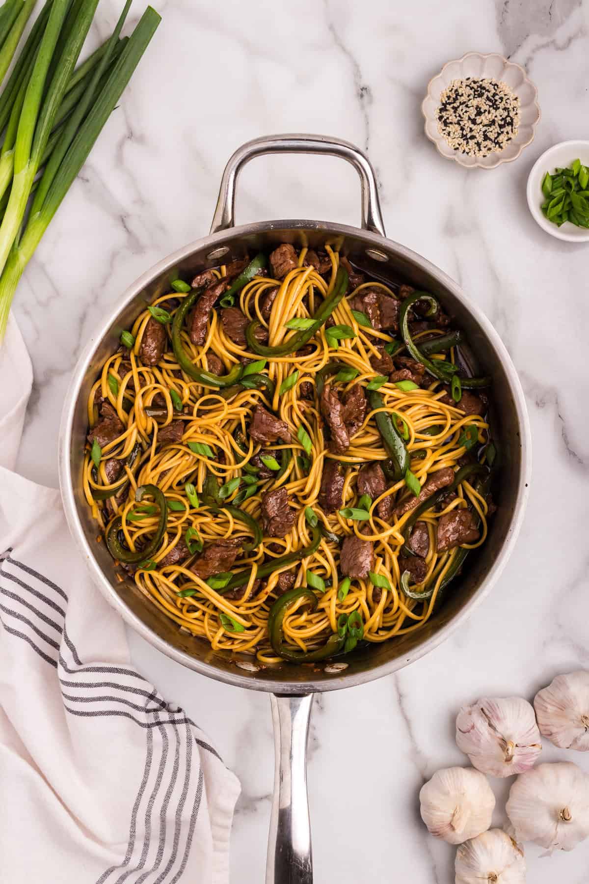 Finished Mongolian beef noodles garnished with chopped green onions and surrounded by garlic, sesame seeds, and scallions on a marble countertop.