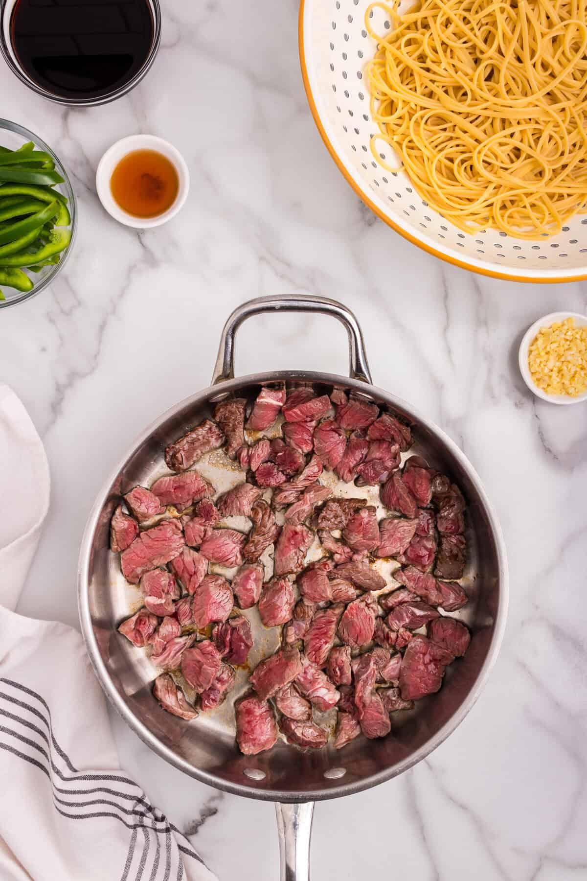 Raw beef slices beginning to brown in a stainless steel skillet; surrounding ingredients include green bell peppers, soy sauce, sesame oil, garlic, and cooked noodles.