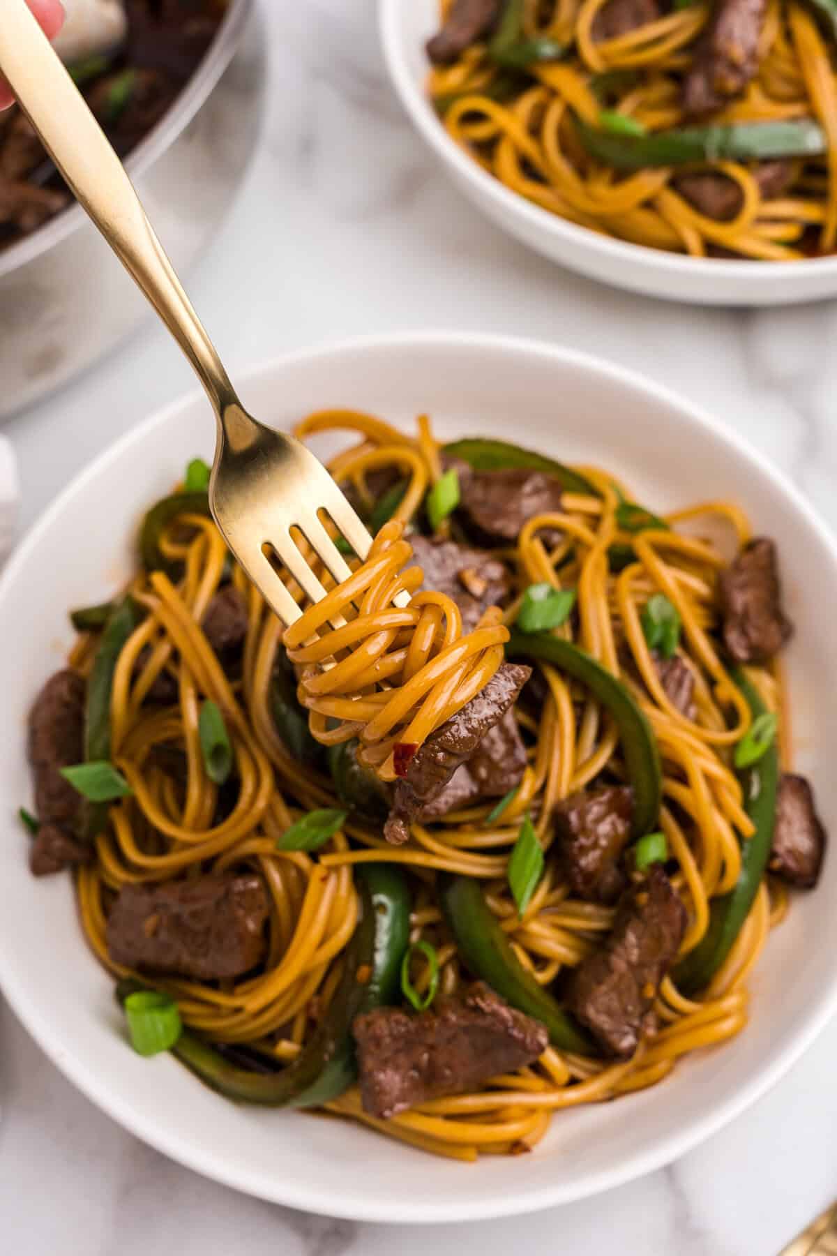 A fork lifting a twirl of Mongolian beef noodles from a serving bowl, with beef and peppers visible below.