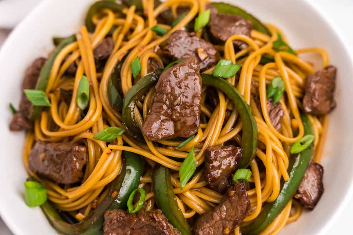 Extreme close-up of Mongolian beef noodles in a white bowl, focusing on the glistening beef slices, noodles, and green onions.