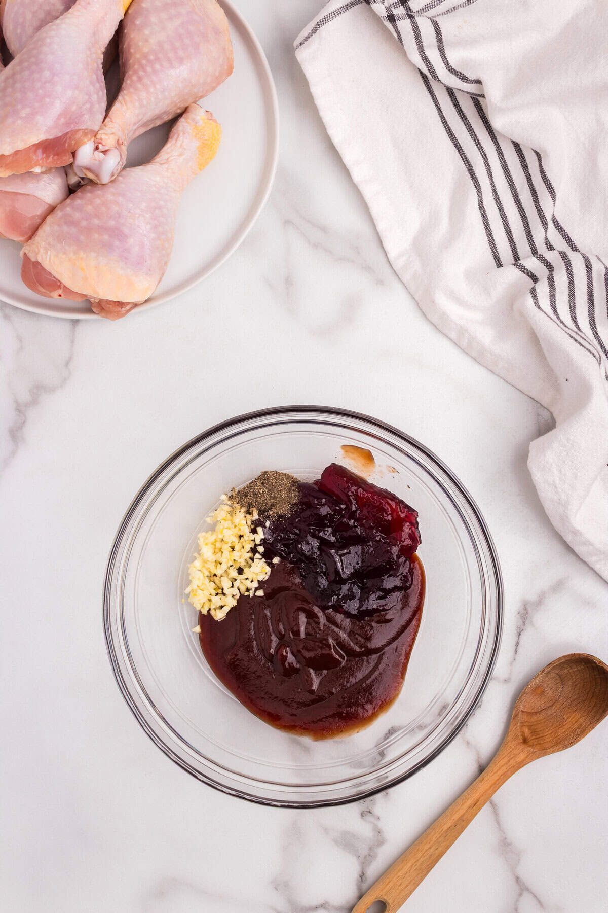 A bowl containing uncombined ingredients for a BBQ marinade—minced garlic, black pepper, BBQ sauce, and what appears to be a fruit preserve or jam, next to a plate of raw chicken drumsticks.
