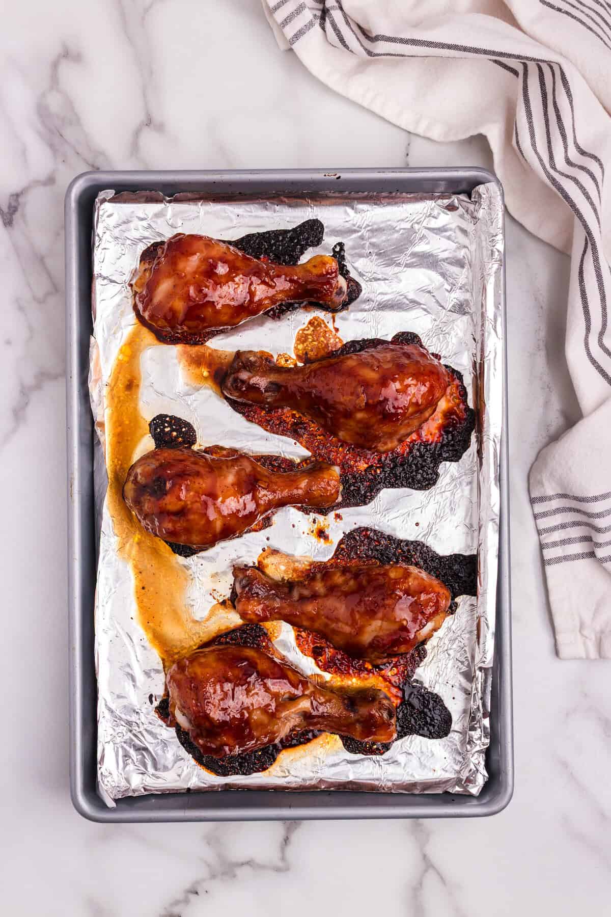 Fully baked BBQ chicken drumsticks on the foil-lined sheet pan, showing caramelization and a glossy, darkened coating.